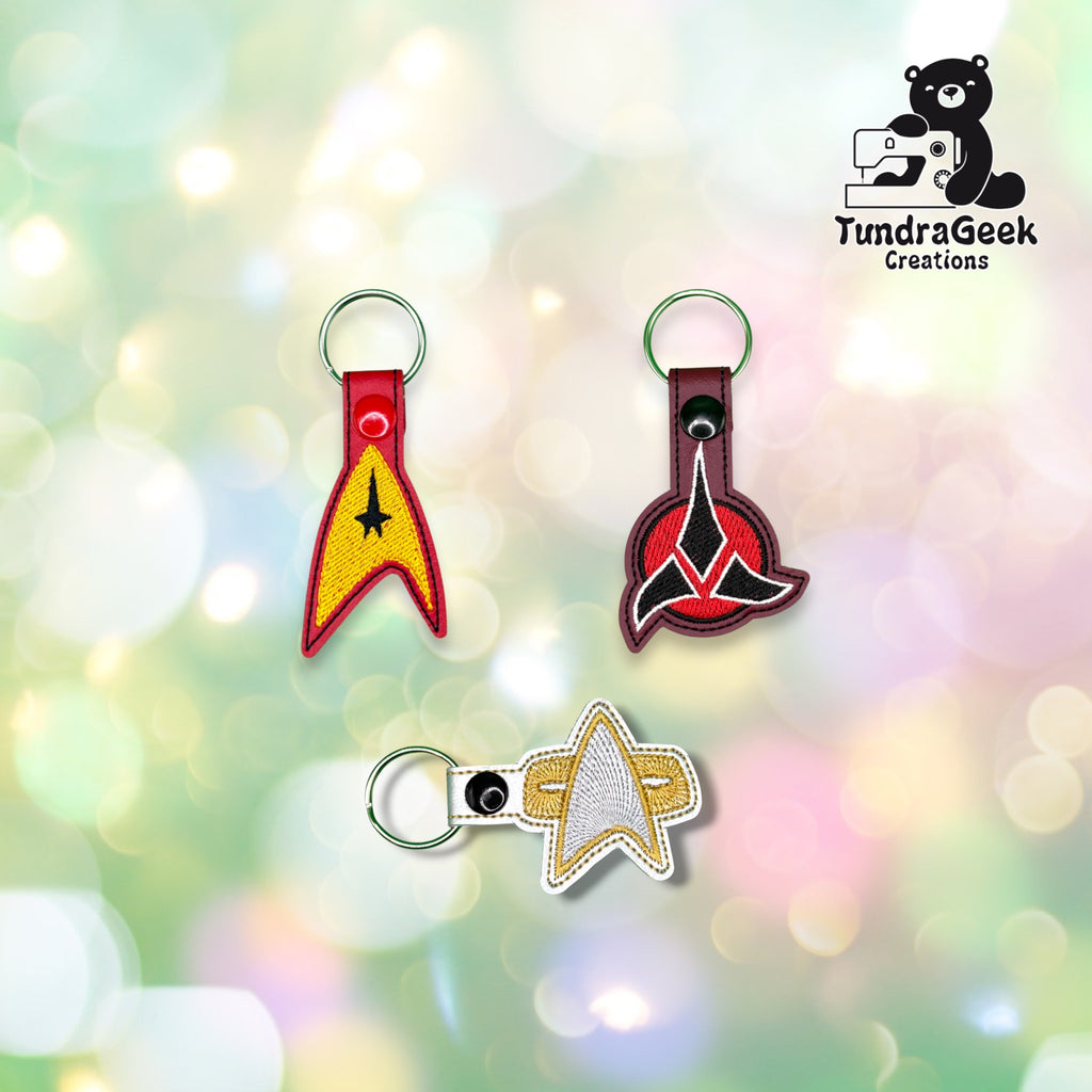 Three embroidered insignia-shaped keychains displayed together on a blurred background. TundraGeek Creations logo in the top right corner.