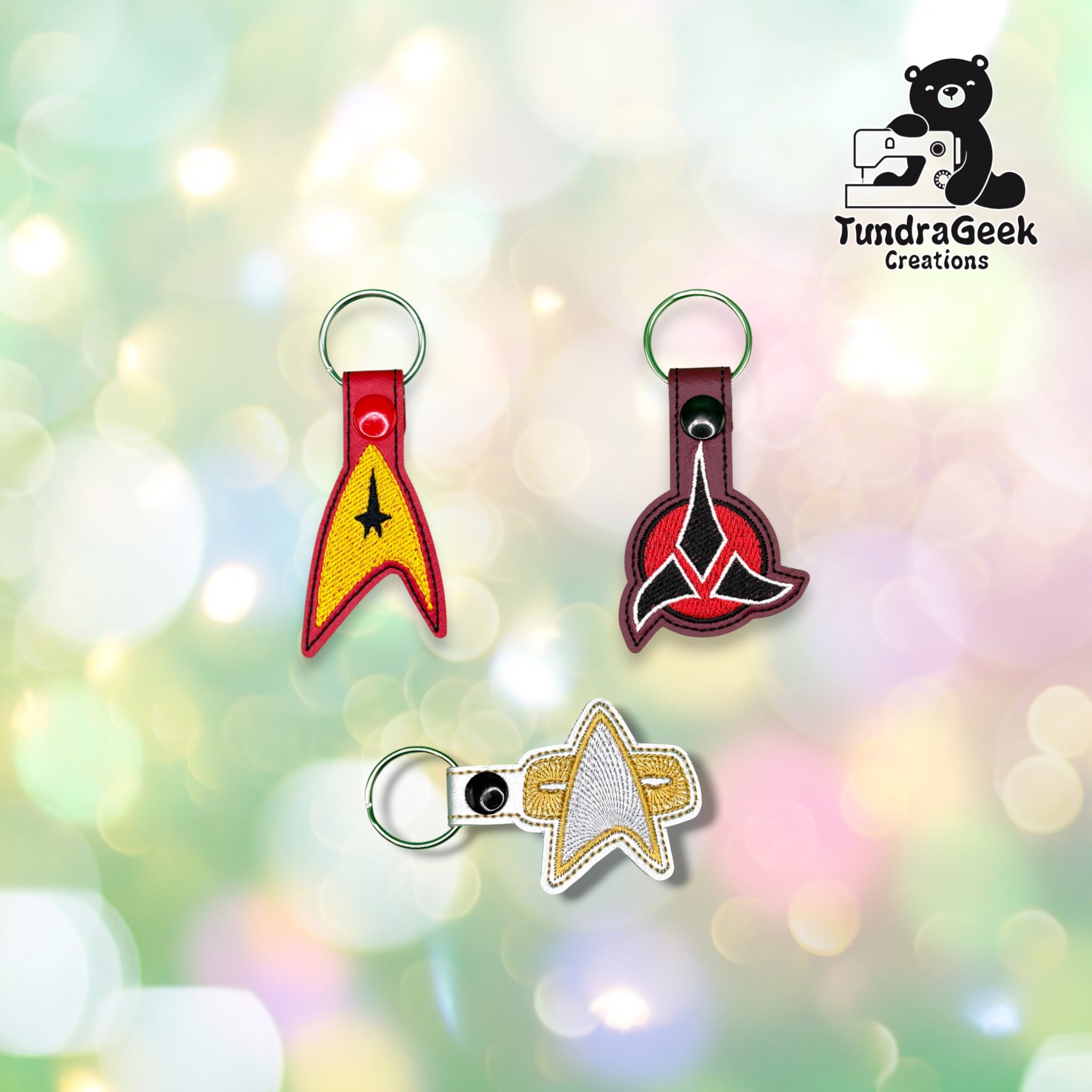 Three embroidered insignia-shaped keychains displayed together on a blurred background. TundraGeek Creations logo in the top right corner.