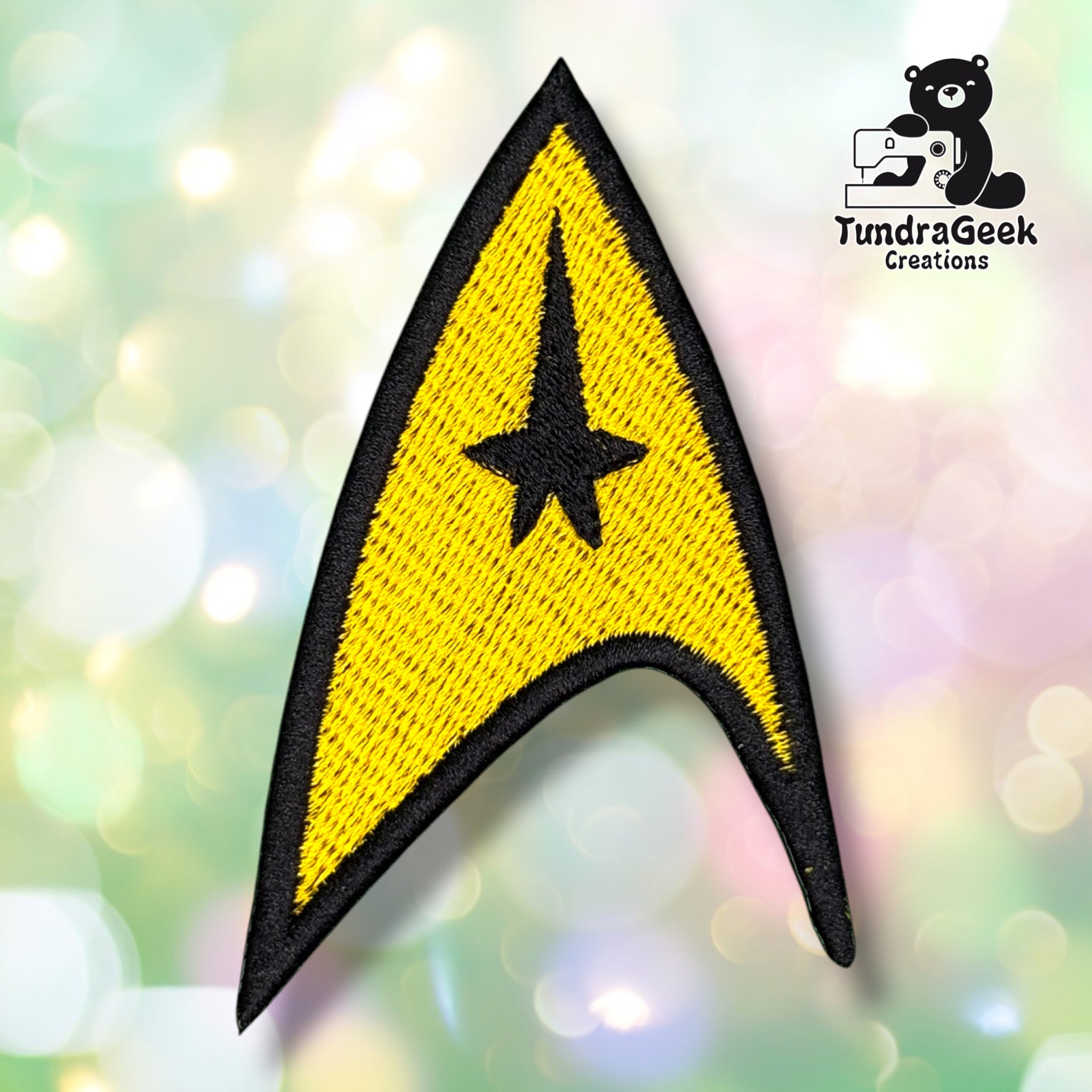 A gold and black arrowhead-shaped embroidered patch with a star motif, on a blurred pastel background. TundraGeek Creations logo in the top right corner.