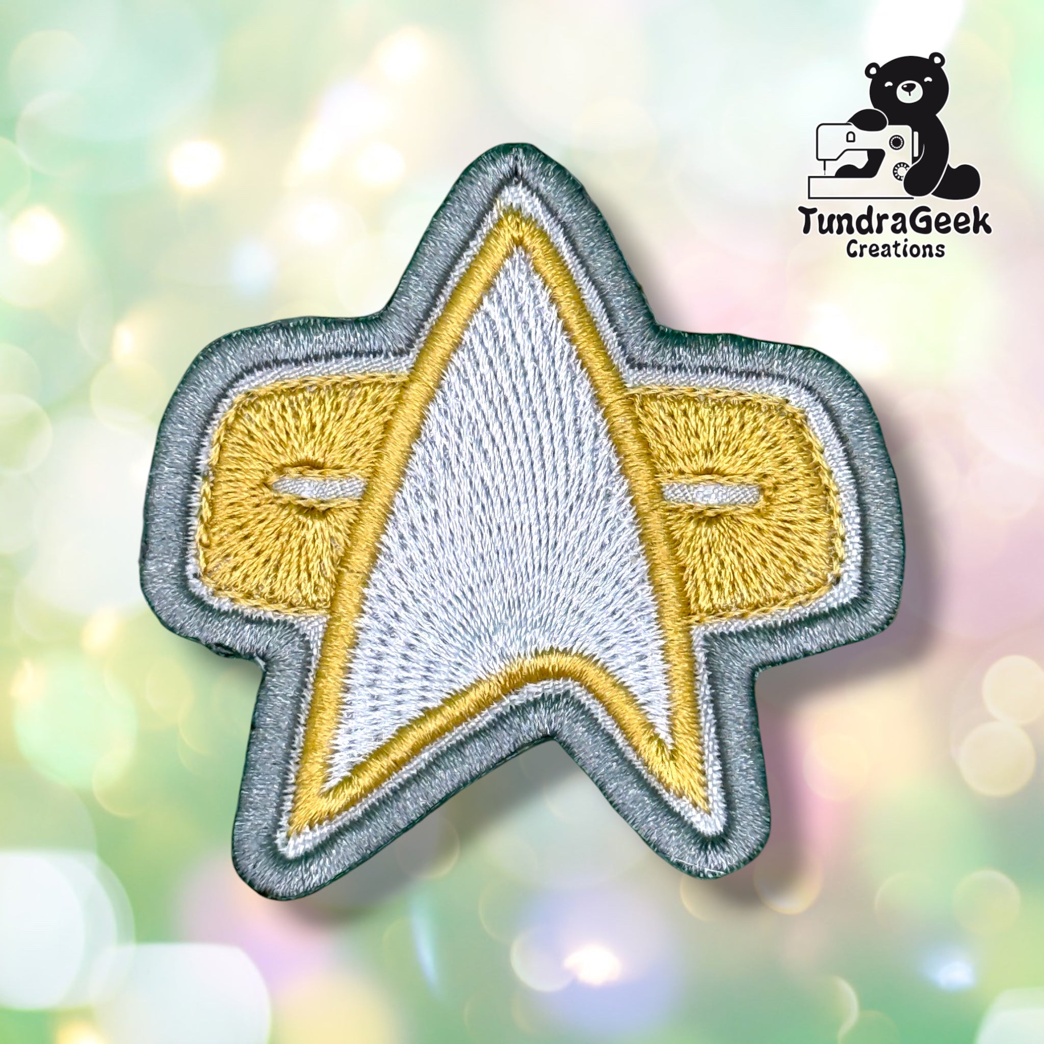 A silver and gold star-shaped embroidered patch with clean detail shot on a blurred pastel background. TundraGeek Creations logo in the top right corner.
