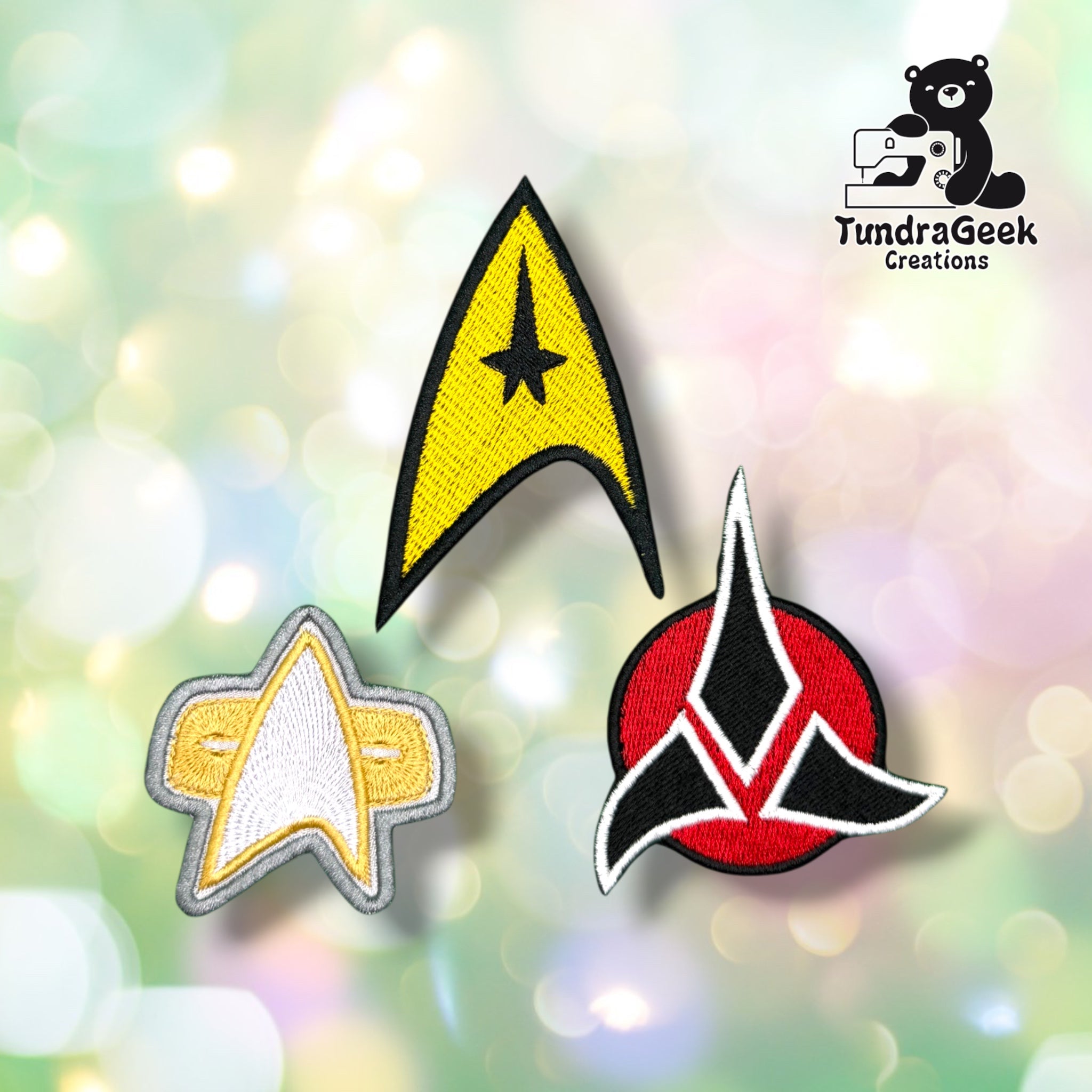 Three embroidered patches — a gold arrowhead, a silver star, and a red crest — displayed together on a blurred pastel background. TundraGeek Creations logo in the top right corner.