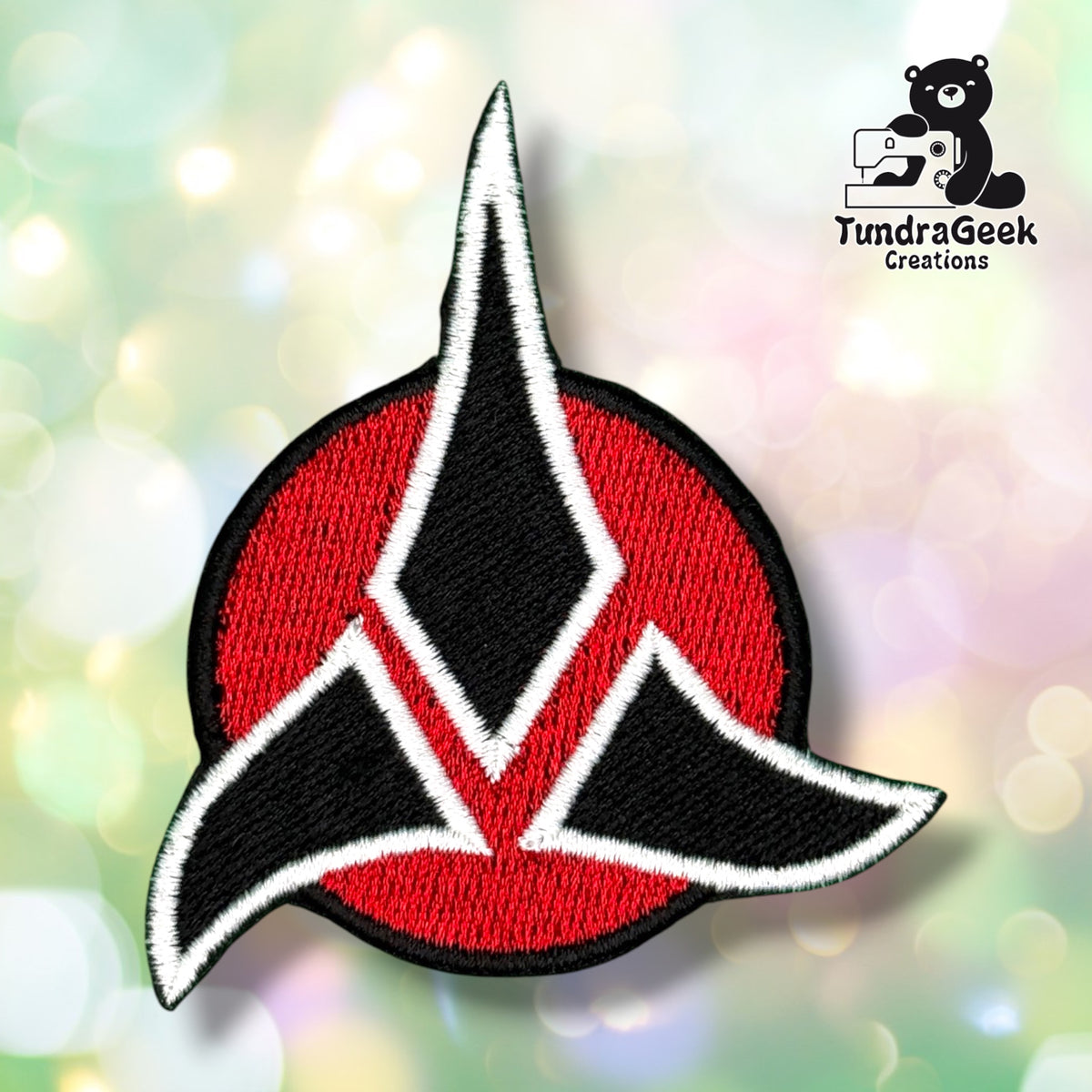 A red and black embroidered crest-shaped patch on a blurred pastel background. TundraGeek Creations logo in the top right corner.