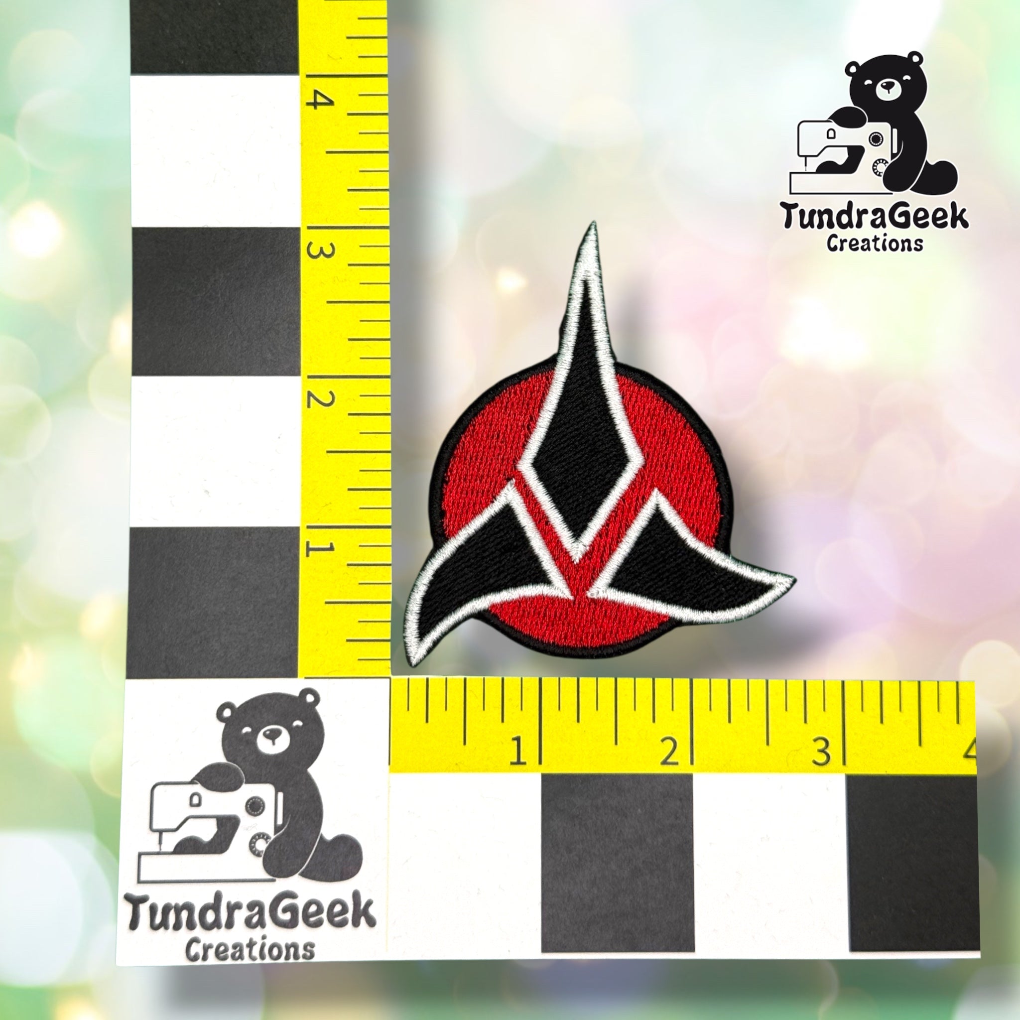 A red and black embroidered crest-shaped patch, shown next to a ruler measuring approximately 3 inches. TundraGeek Creations logo in the top right corner.