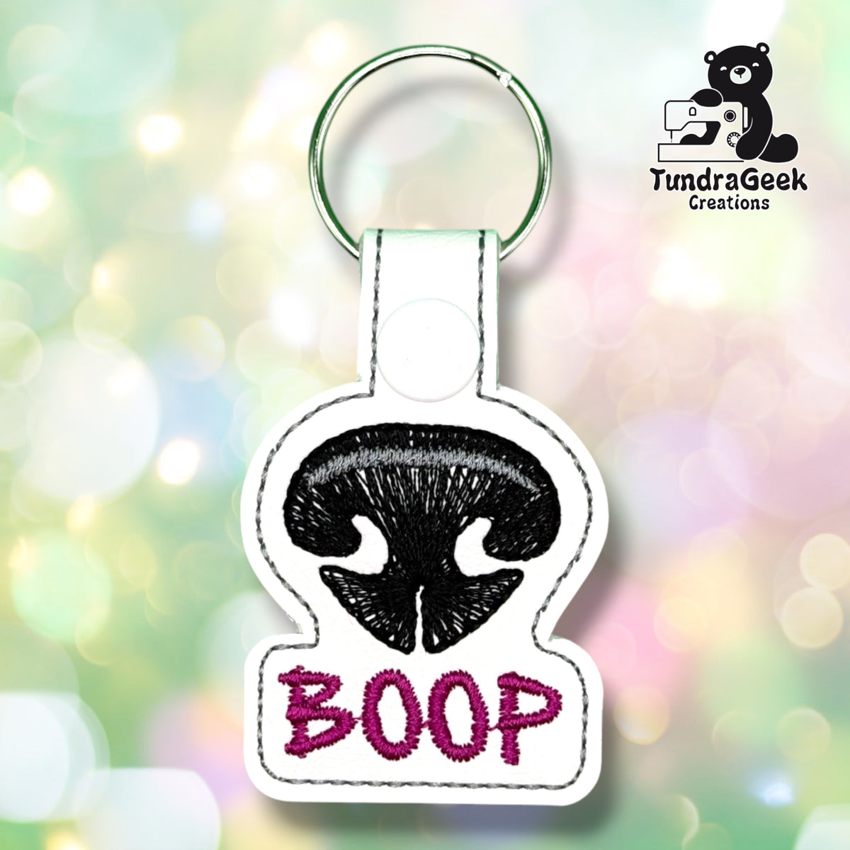 Front view of a white vinyl keychain embroidered with a black dog nose featuring a grey highlight stripe, with "BOOP" stitched below in magenta lettering. Finished with a white plastic snap closure and green split ring.