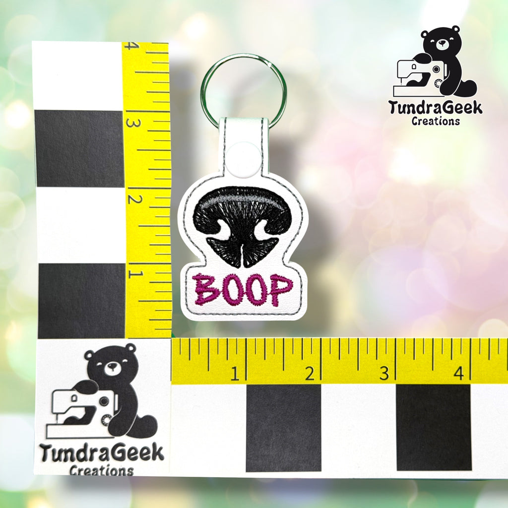 Keychain shown against a black-and-white checkered background with yellow rulers for scale, measuring approximately 1.75 inches wide by 3.75 inches tall, including hardware.  ---