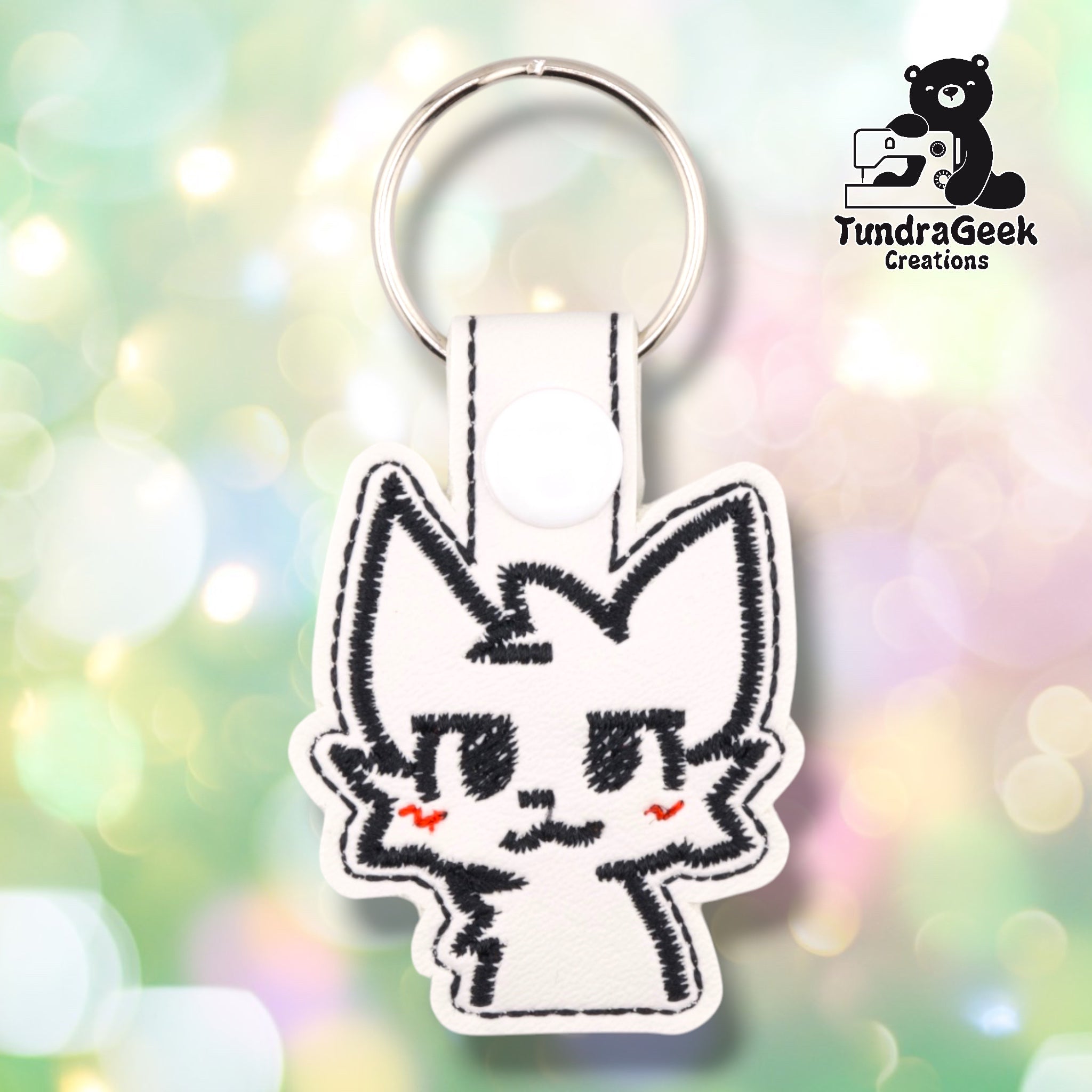 Front view of a white faux leather keychain shaped like a stylized cat face, with black stitched outlines for the ears, eyes, and mouth, and a red zigzag stitch on each cheek suggesting a flustered blush. A white 12.5mm plastic snap closure secures the tab at the top, with a silver split ring attached above.