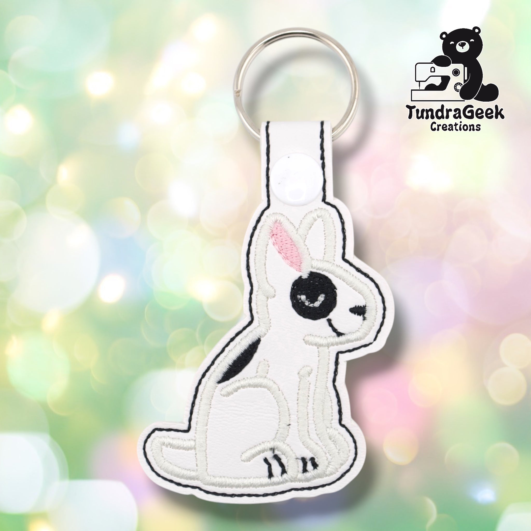 White vinyl keychain die-cut in the shape of a seated bull terrier. The dog is embroidered in black and white with a distinctive black eye patch, pink inner ear detail, small black nose, and black toe details at the feet. A white snap tab and silver split ring are attached at the top.
