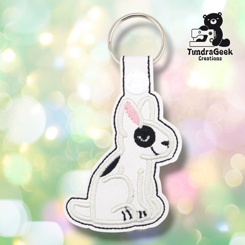 White vinyl keychain die-cut in the shape of a seated bull terrier. The dog is embroidered in black and white with a distinctive black eye patch, pink inner ear detail, small black nose, and black toe details at the feet. A white snap tab and silver split ring are attached at the top.