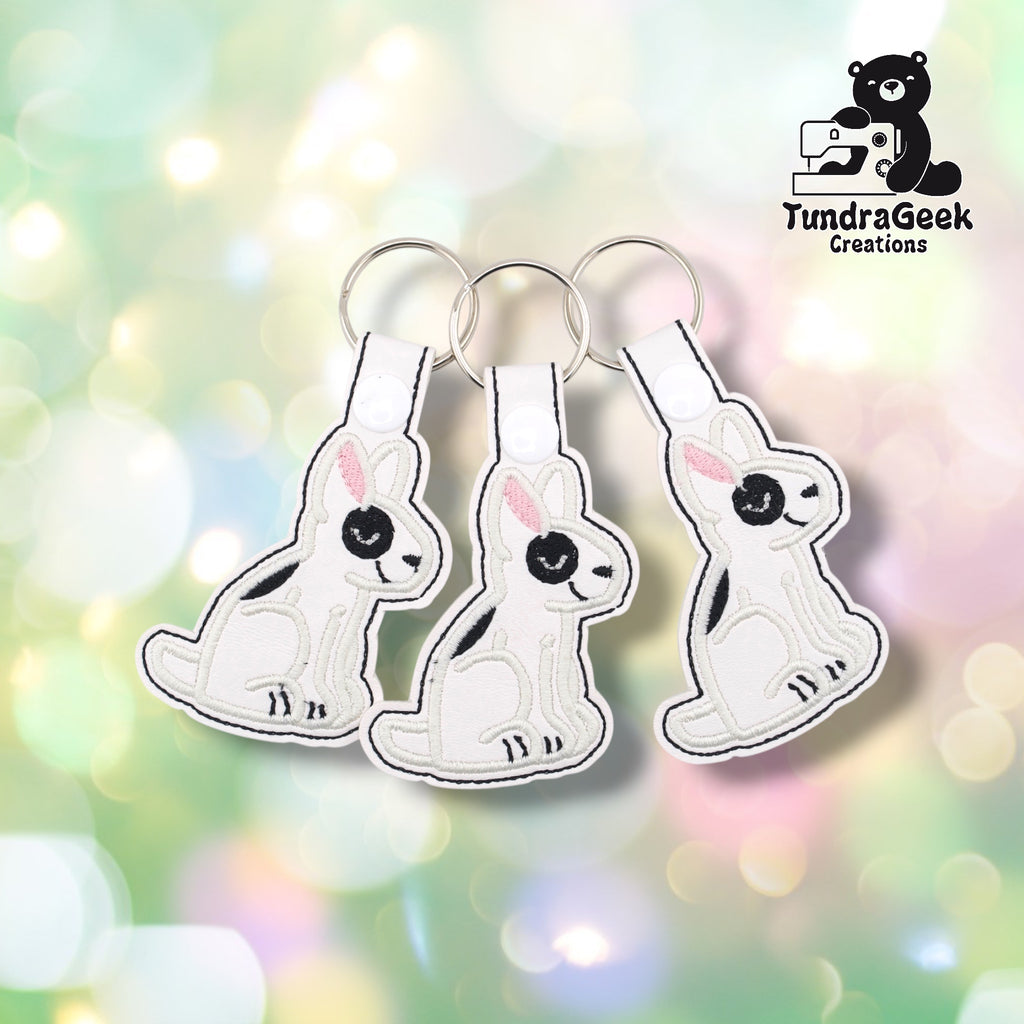 Three bull terrier keychains fanned out on a soft bokeh background, showing the same design from slightly different angles. Each features a white vinyl seated bull terrier with black embroidered markings, black eye patch, and pink ear detail. Silver split ring hardware on each.