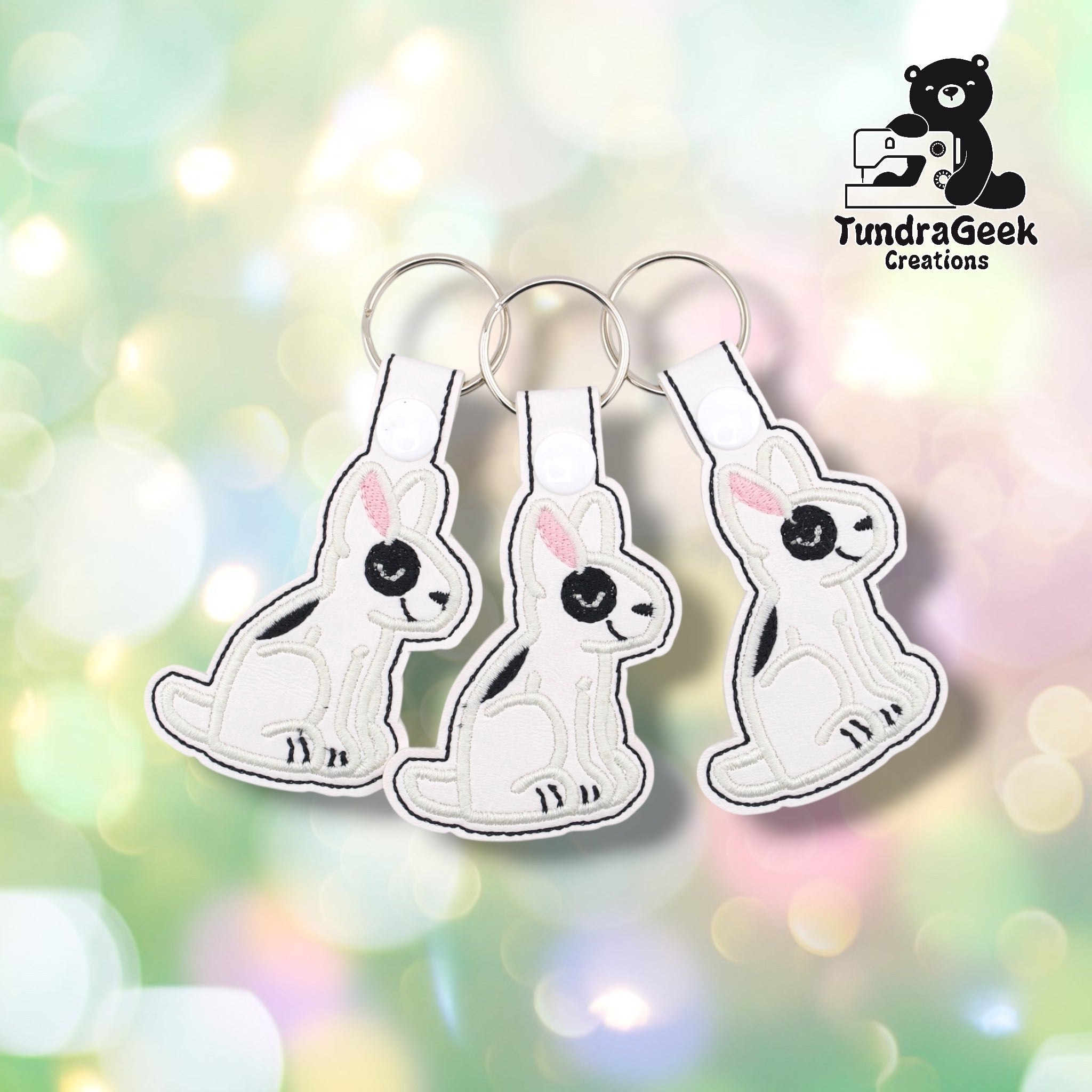 Three bull terrier keychains fanned out on a soft bokeh background, showing the same design from slightly different angles. Each features a white vinyl seated bull terrier with black embroidered markings, black eye patch, and pink ear detail. Silver split ring hardware on each.