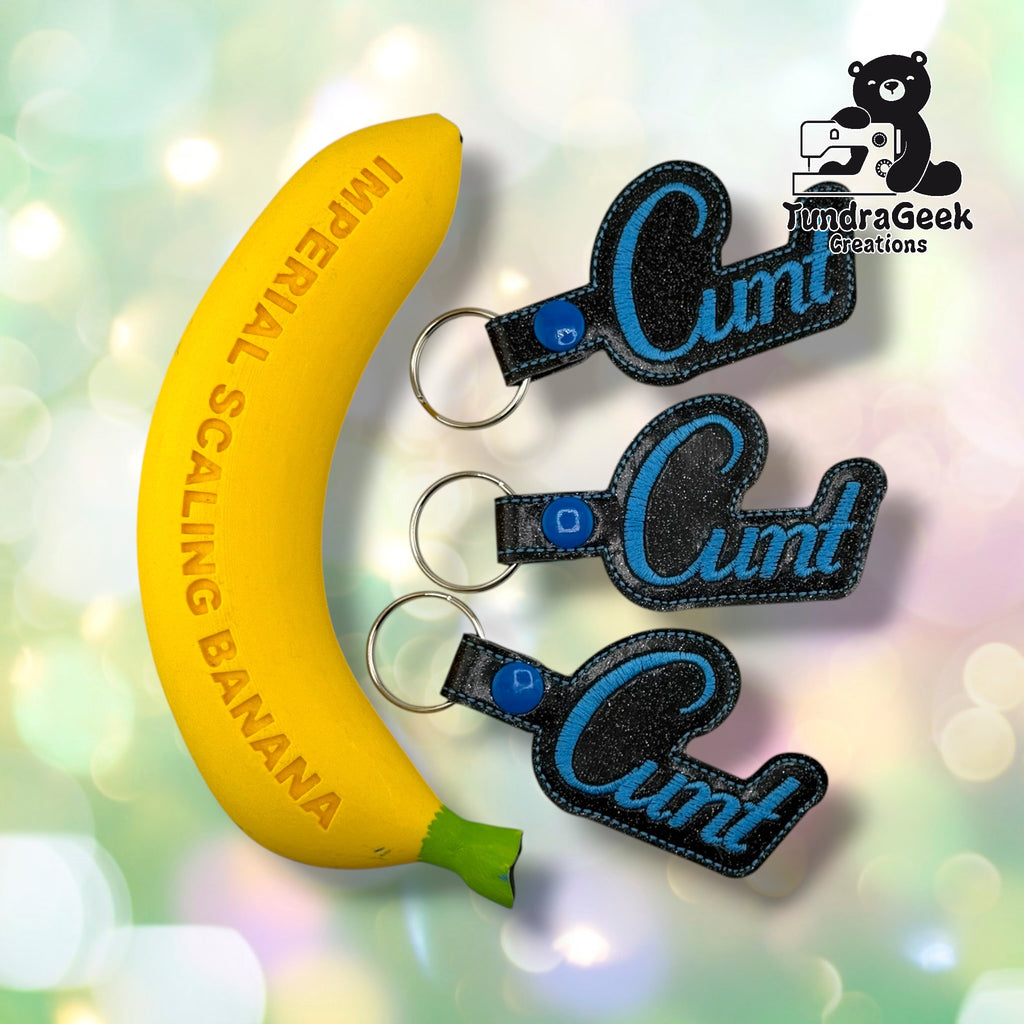 Three black embroidered keychains with blue sparkle vinyl lettering spelling "Cunt," displayed next to a yellow banana-shaped prop for scale. TundraGeek Creations logo visible in top right corner.