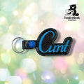 Black sparkle vinyl keychain shaped like a thumbs up with blue embroidered text reading "Cunt," attached to a metal keyring. TundraGeek Creations logo visible in top right corner.
