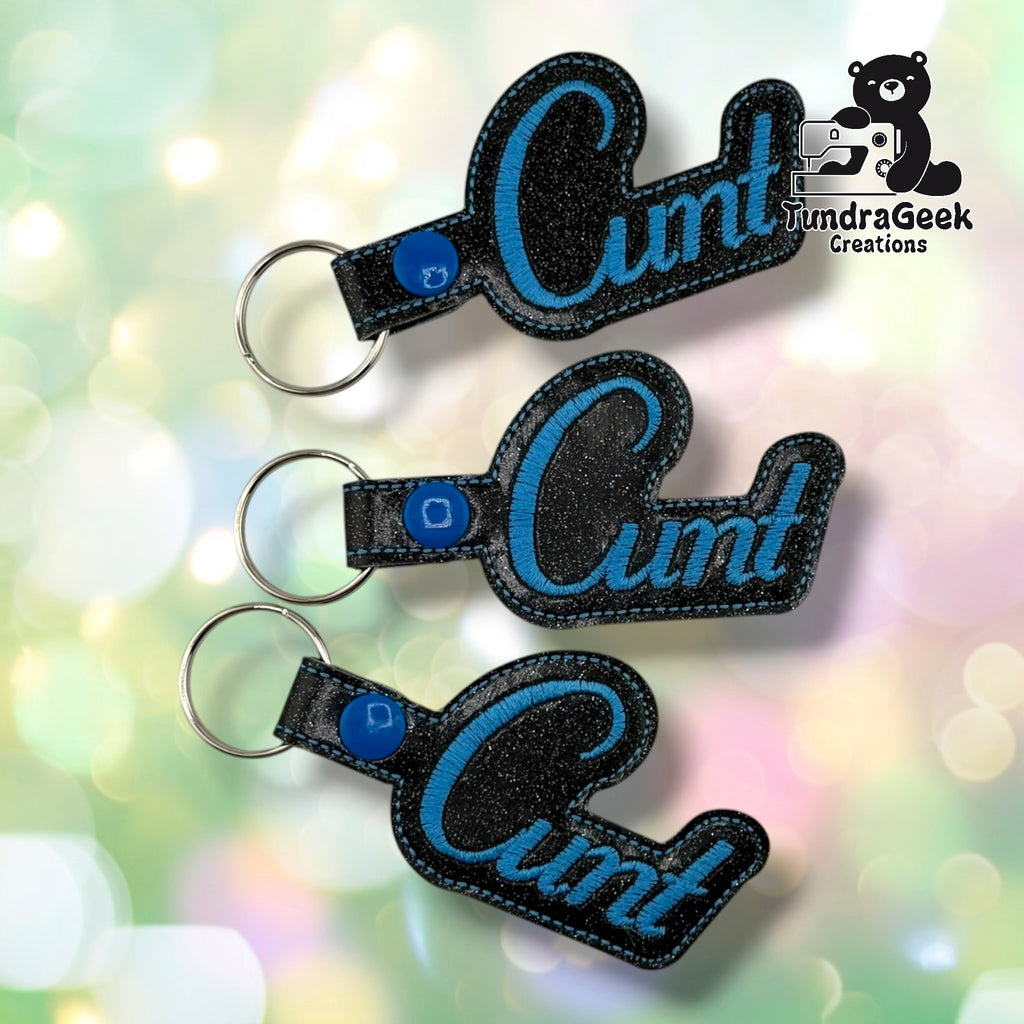 Three black embroidered keychains with blue sparkle vinyl lettering spelling "Cunt," each attached to a metal keyring. TundraGeek Creations logo visible in top right corner.