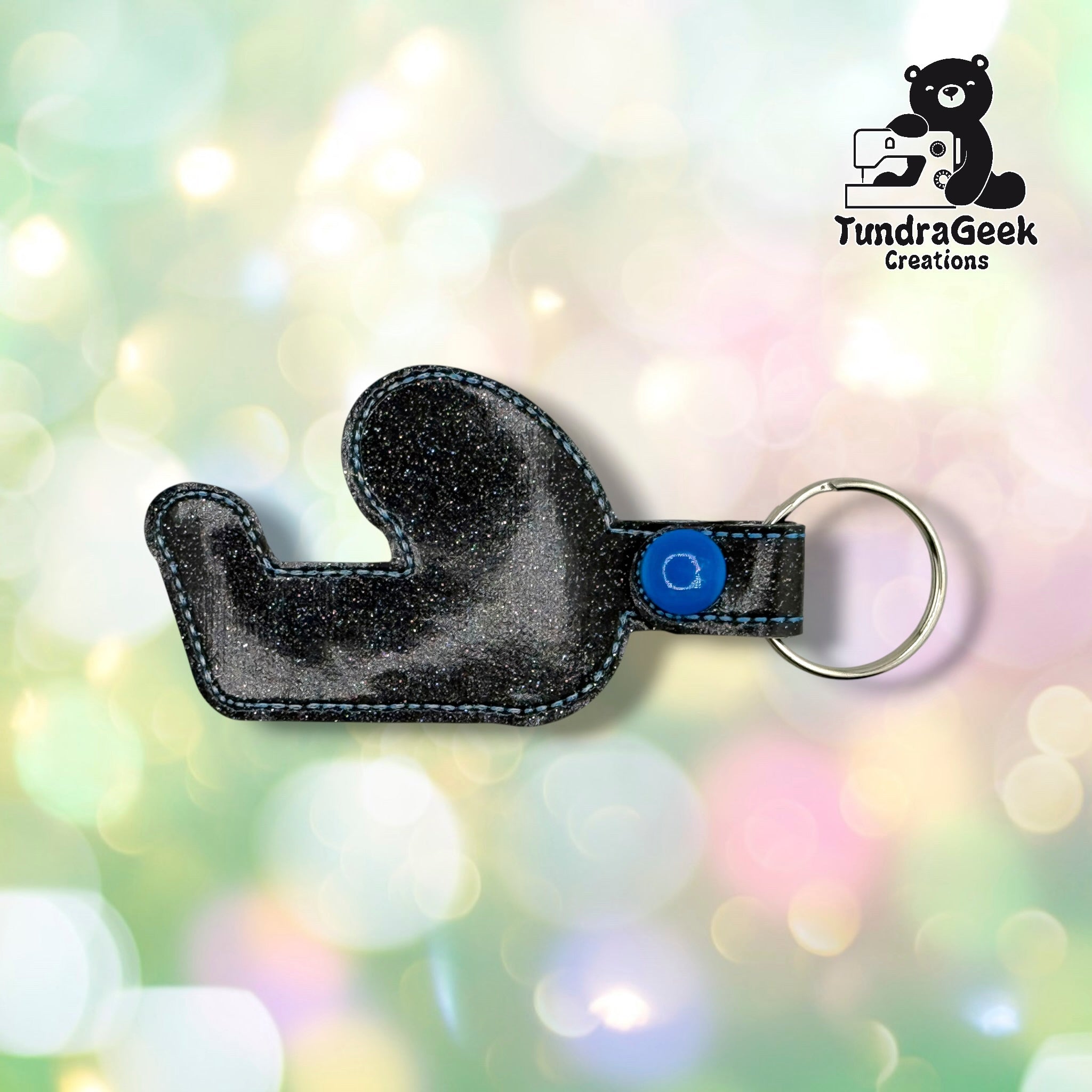 Black sparkle vinyl keychain shaped like a mitten, attached to a blue button and metal keyring. TundraGeek Creations logo visible in top right corner.