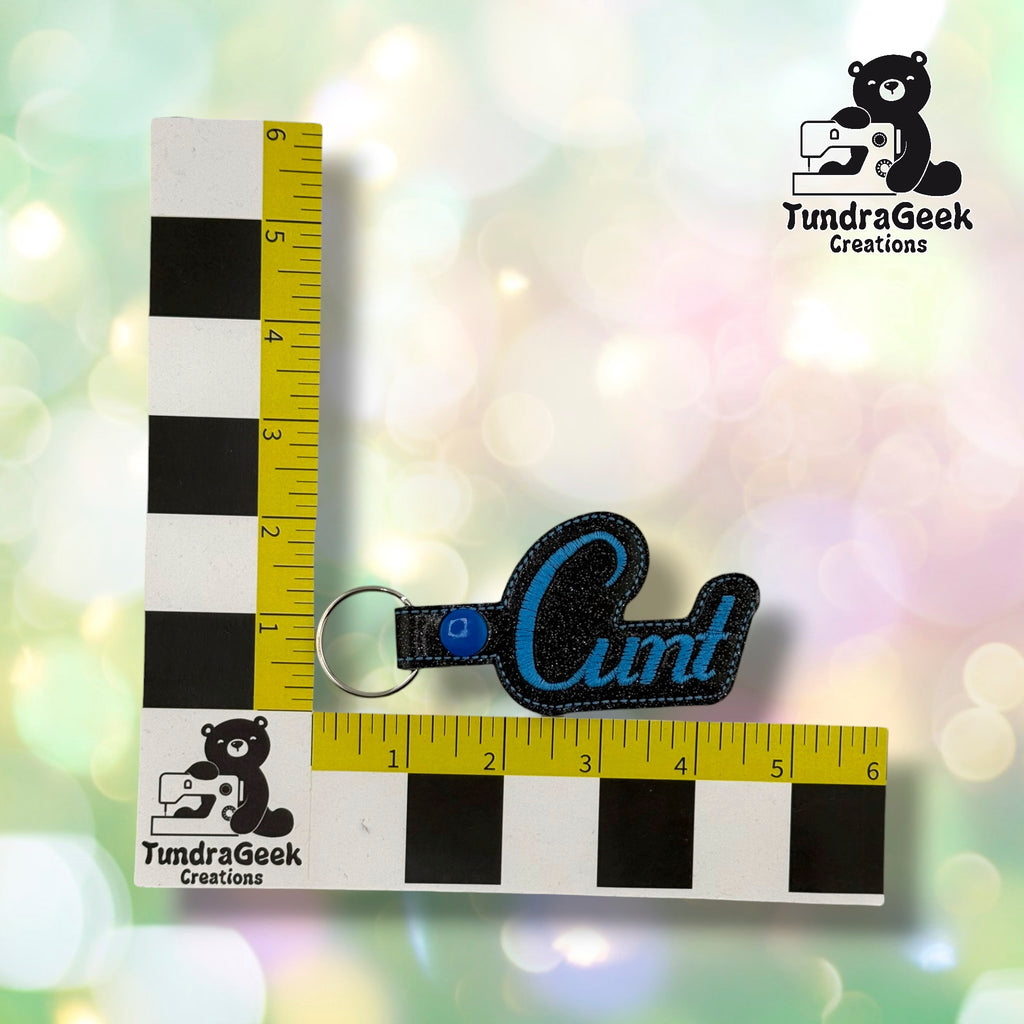 Black sparkle vinyl keychain with blue script text, attached to metal keyring, displayed next to TundraGeek Creations measurement scale.