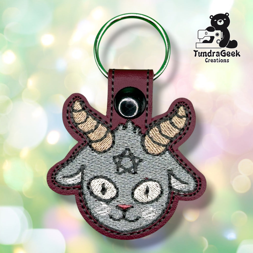 Front view of a maroon vinyl keychain embroidered with a grey Baphomet goat face, featuring striped tan horns, cat-like eyes, and a small star between the brows. Finished with a black plastic snap closure and green split ring.