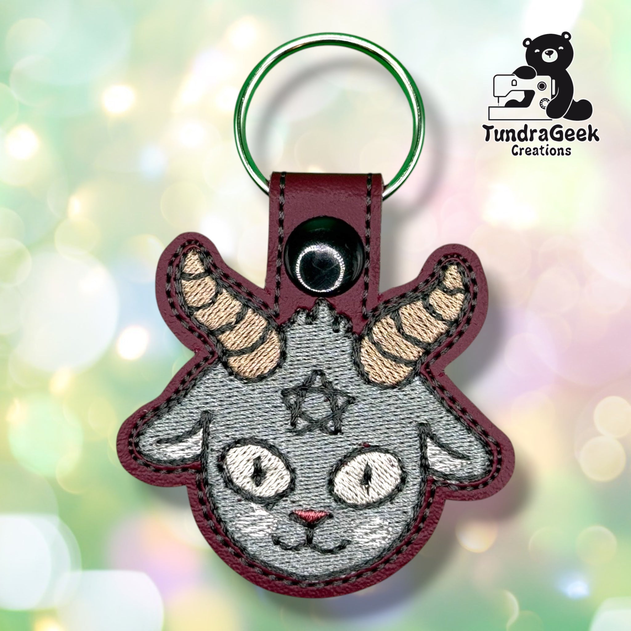 Front view of a maroon vinyl keychain embroidered with a grey Baphomet goat face, featuring striped tan horns, cat-like eyes, and a small star between the brows. Finished with a black plastic snap closure and green split ring.