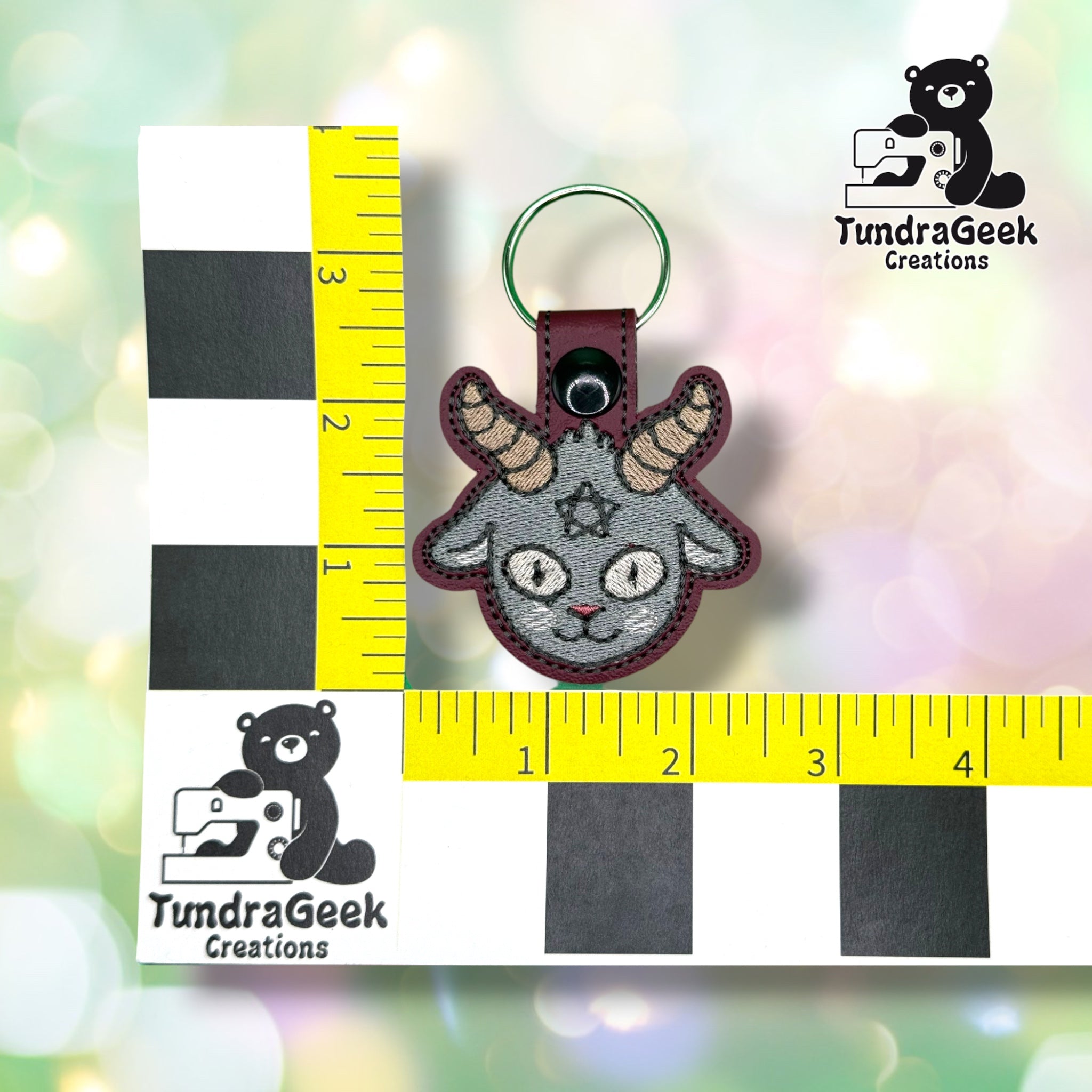 Keychain shown against a black-and-white checkered background with yellow rulers for scale, measuring approximately 2.5 inches wide by 3.5 inches tall, including hardware.  ---