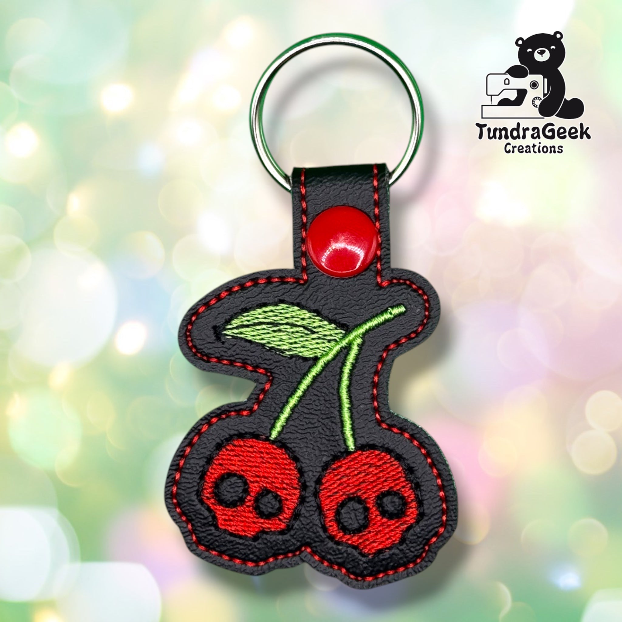 Front view of a black vinyl keychain featuring two red cherries with black skull faces embroidered in place of the pits, joined by green stems and a single green leaf, finished with red stitched edge detailing and a red plastic snap closure.