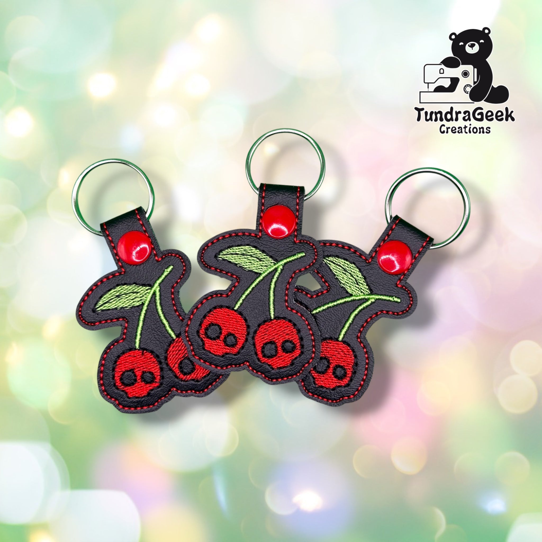 Three of the same keychain shown together, each with two red skull-faced cherries, green stems and a leaf, black vinyl backing, red plastic snap closures, and green split rings.