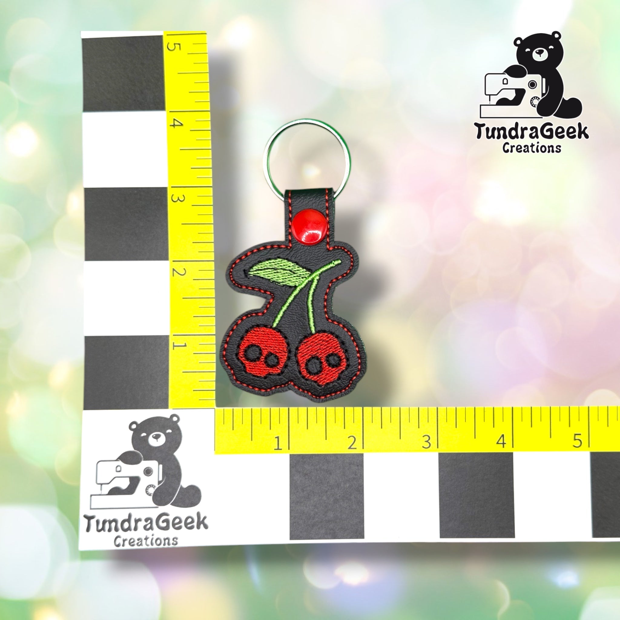 Keychain shown against a black-and-white checkered background with yellow rulers for scale, measuring approximately 4 inches tall by 2 inches wide including hardware.  ---