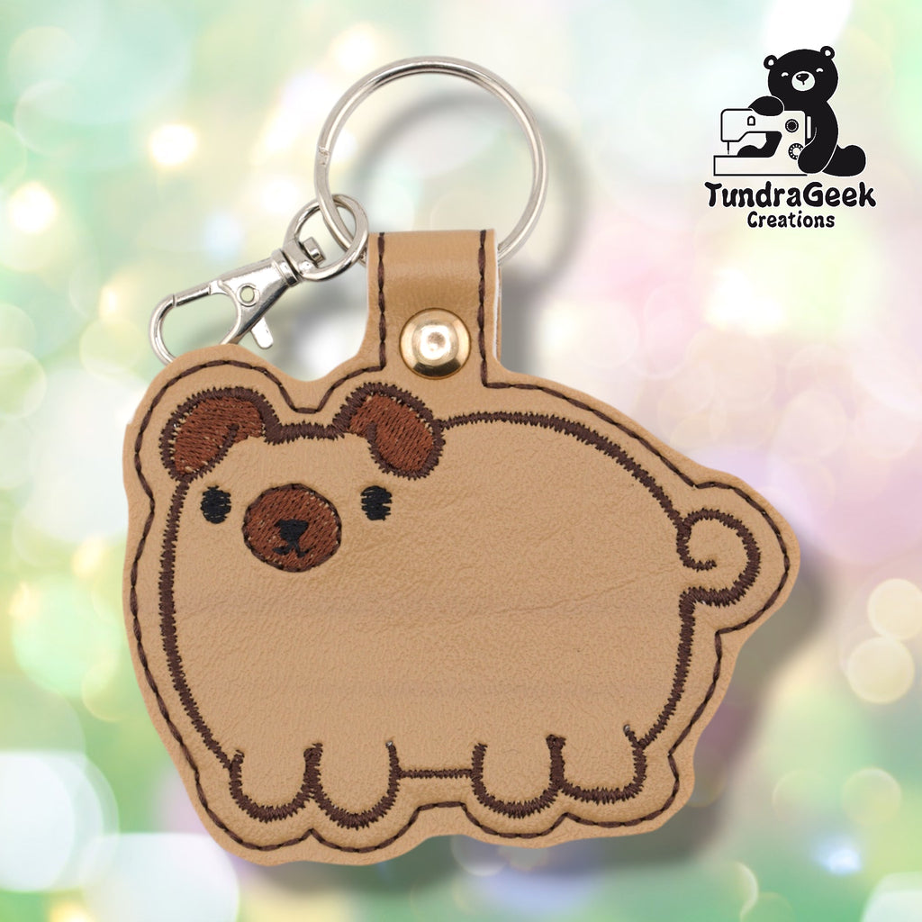 Tan vinyl keychain die-cut in the shape of a chubby, round pug. The dog is embroidered with brown ear markings, a brown face mask, small black eyes, a brown nose, and a curly tail detail on the right side. Four stubby legs are visible along the bottom edge. Gold snap hardware and silver lobster claw clip with split ring at top.