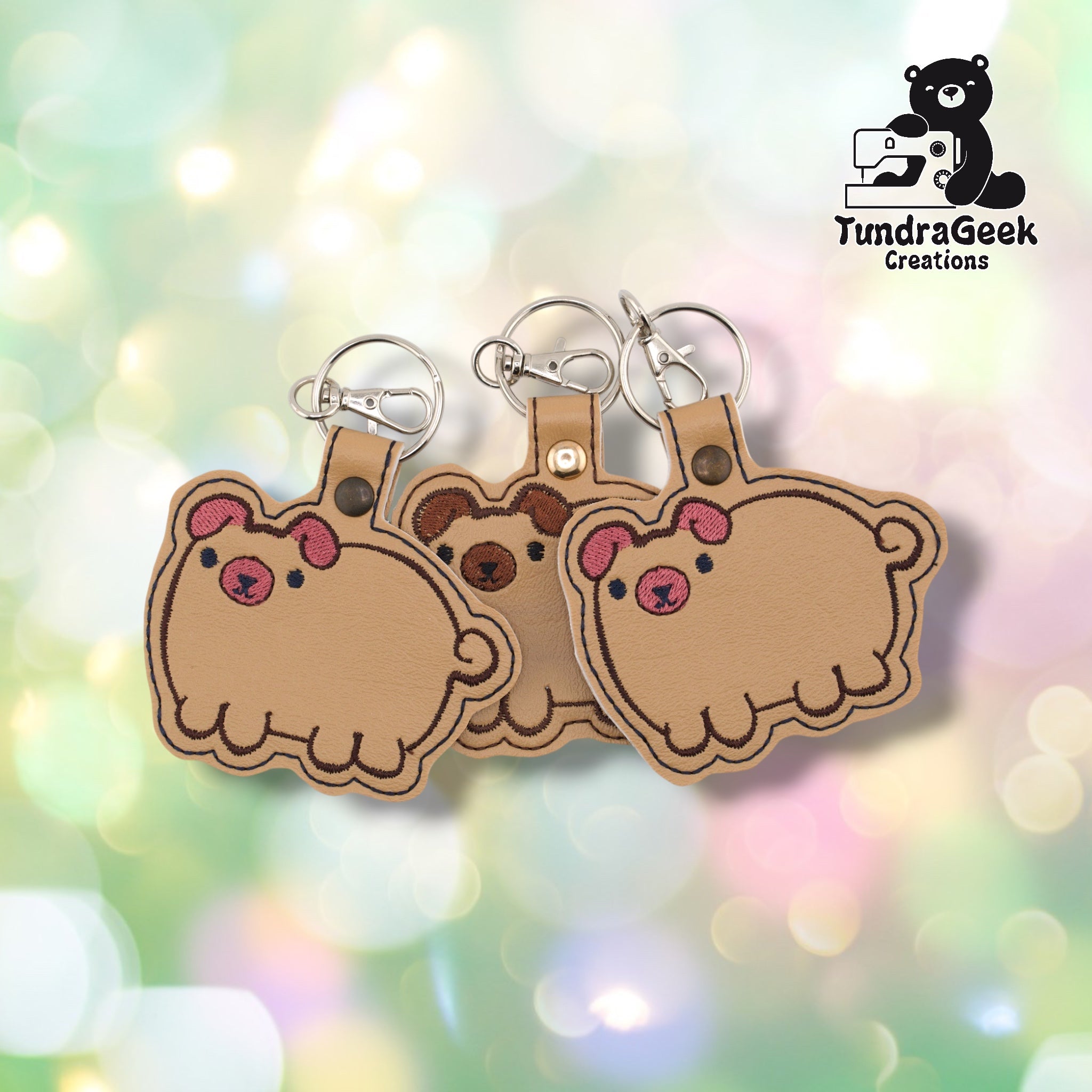 Three chubby pug keychains fanned out on a soft bokeh background. Left and right keychains show pink ear and face markings; the center keychain shows brown markings. All three are on tan vinyl with silver lobster claw clip and split ring hardware. The round, wide shape of the pug body is visible across all three.