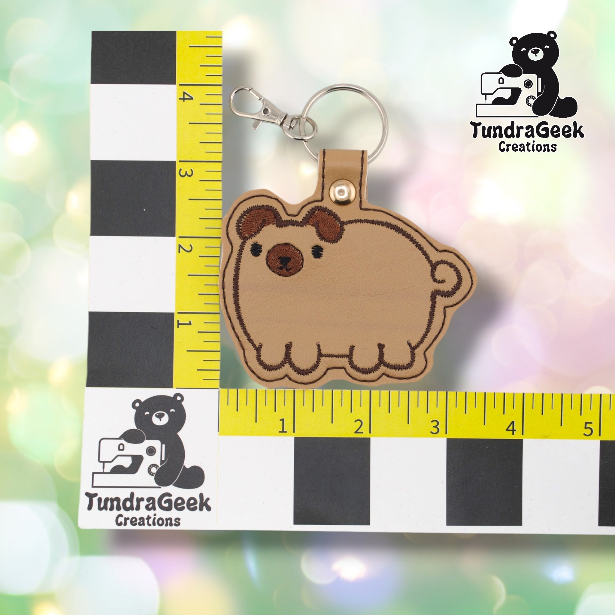 Chubby pug keychain shown against a black-and-white checkered background with yellow rulers for scale. The keychain body measures approximately 3.5 inches wide by 3.25 inches tall, not including hardware. Brown marking variant shown. Gold snap hardware and silver lobster claw clip at top.