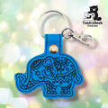 Bright blue vinyl keychain die-cut in the shape of a side-profile elephant with trunk curled downward. Dense black embroidery covers the body with mandala-style circular patterns, six-pointed stars, scrollwork, and floral details across the head, body, and legs. Gold snap hardware and silver lobster claw clip with split ring at top.