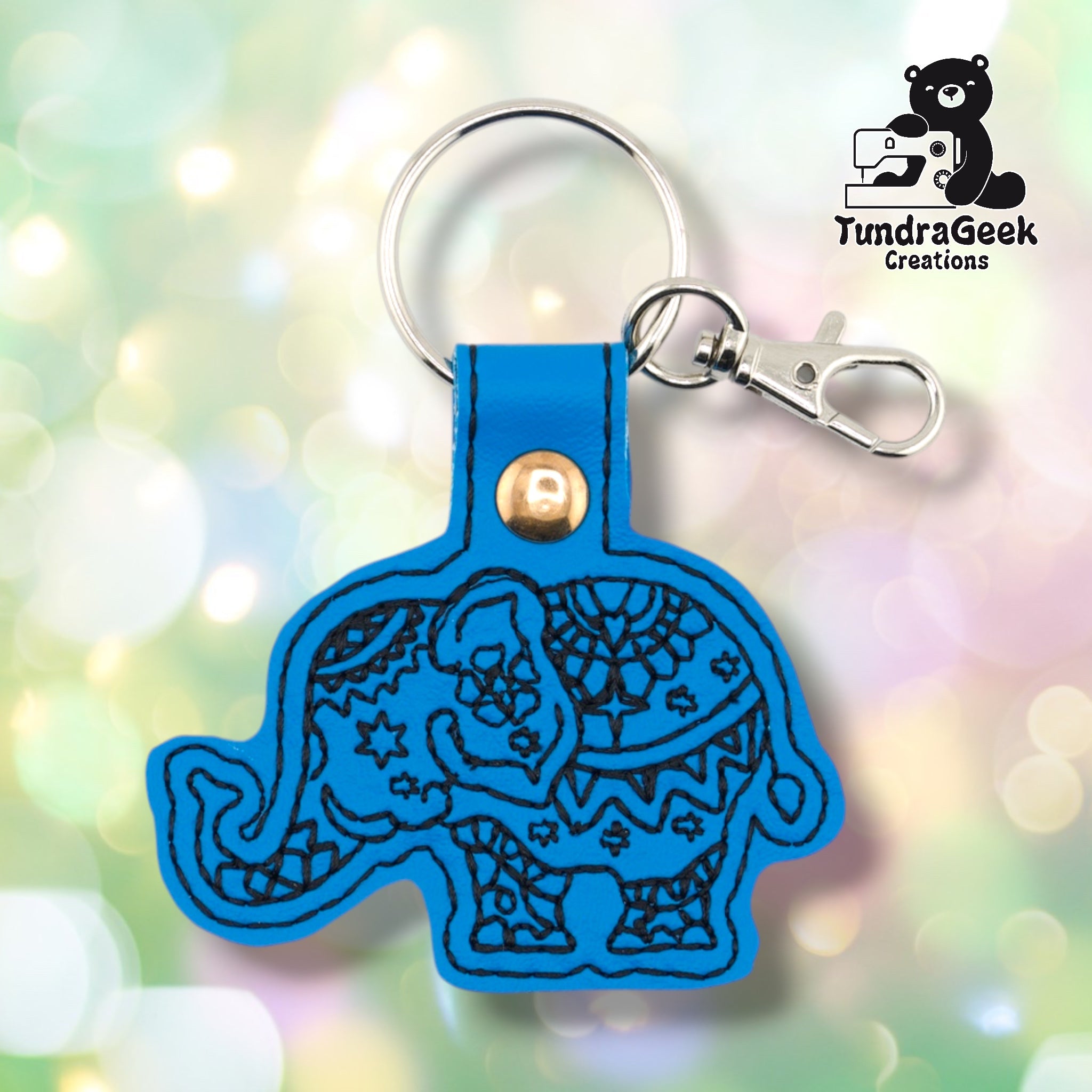 Bright blue vinyl keychain die-cut in the shape of a side-profile elephant with trunk curled downward. Dense black embroidery covers the body with mandala-style circular patterns, six-pointed stars, scrollwork, and floral details across the head, body, and legs. Gold snap hardware and silver lobster claw clip with split ring at top.