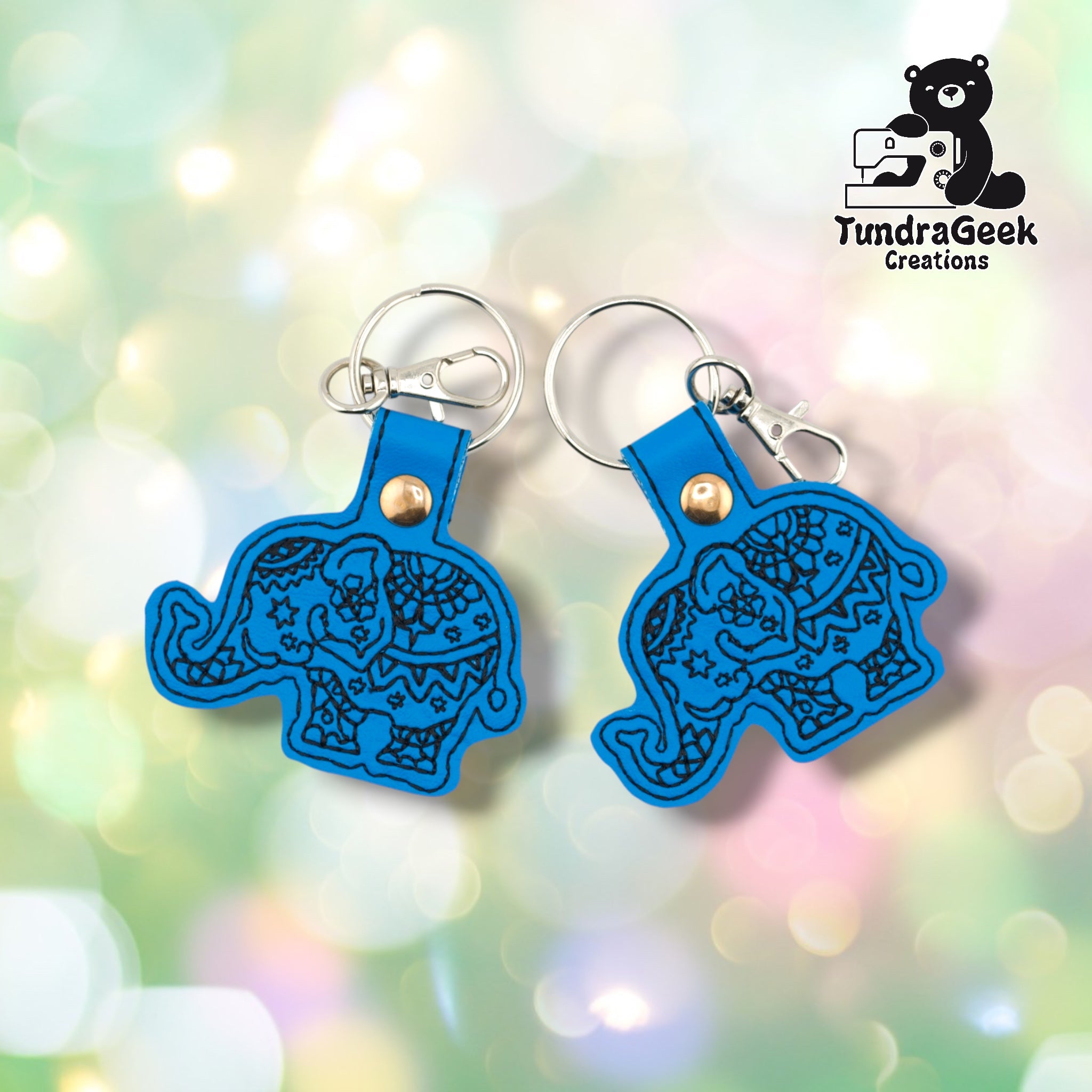 Two circus elephant keychains side by side on a soft bokeh background, showing the same design from slightly different angles. Both feature bright blue vinyl with intricate black embroidered mandala and star patterning across the elephant's body. Gold snap hardware and silver lobster claw clip with split ring on each.
