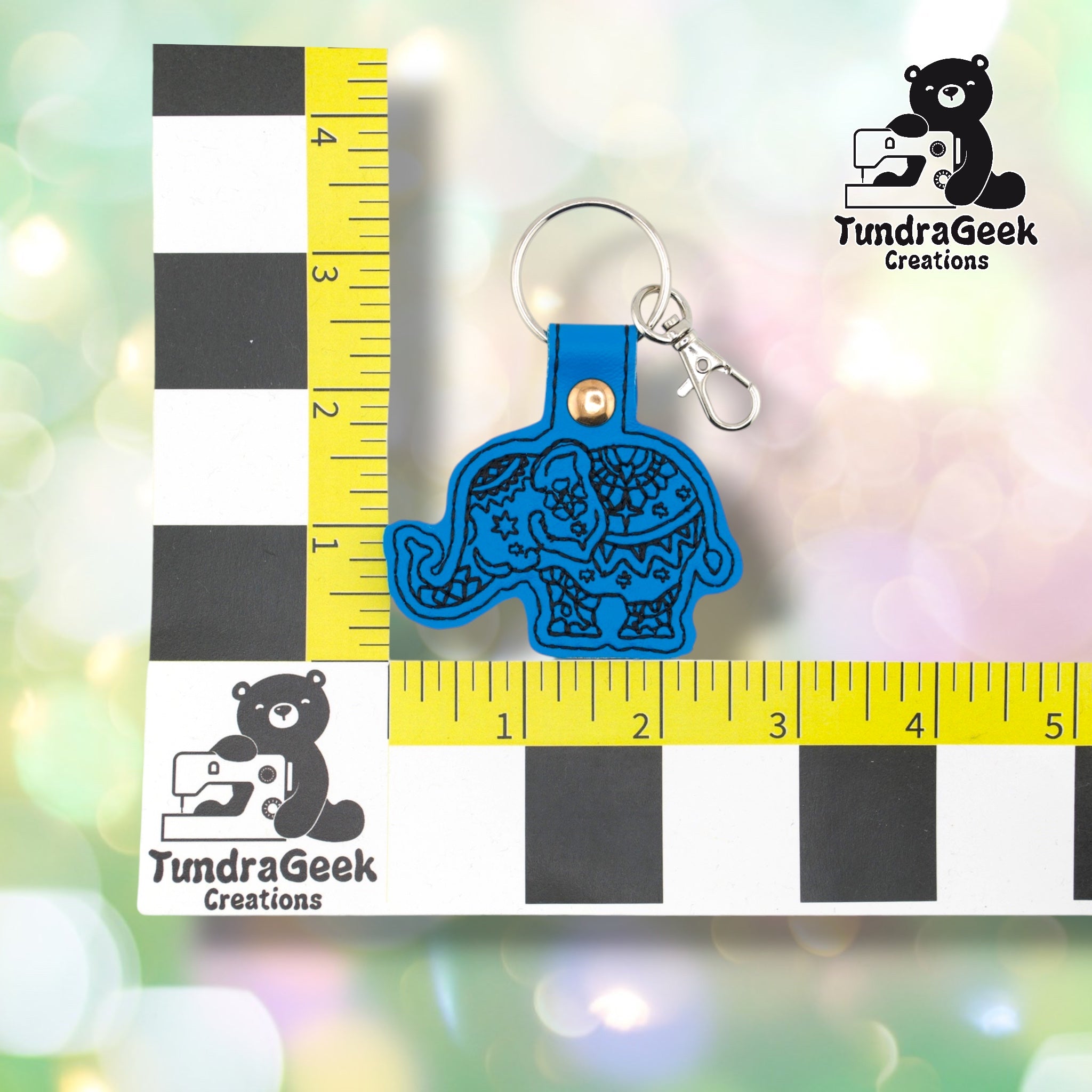 Circus elephant keychain shown against a black-and-white checkered background with yellow rulers for scale. The keychain body measures approximately 2.75 inches wide by 2.5 inches tall, not including hardware. Gold snap hardware and silver lobster claw clip at top.