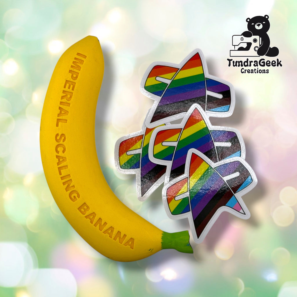 Three identical die-cut vinyl stickers in a delta badge shape filled with Progress Pride flag colors, shown beside the Imperial Scaling Banana, TundraGeek Creations' branded novelty ruler prop, for size reference.