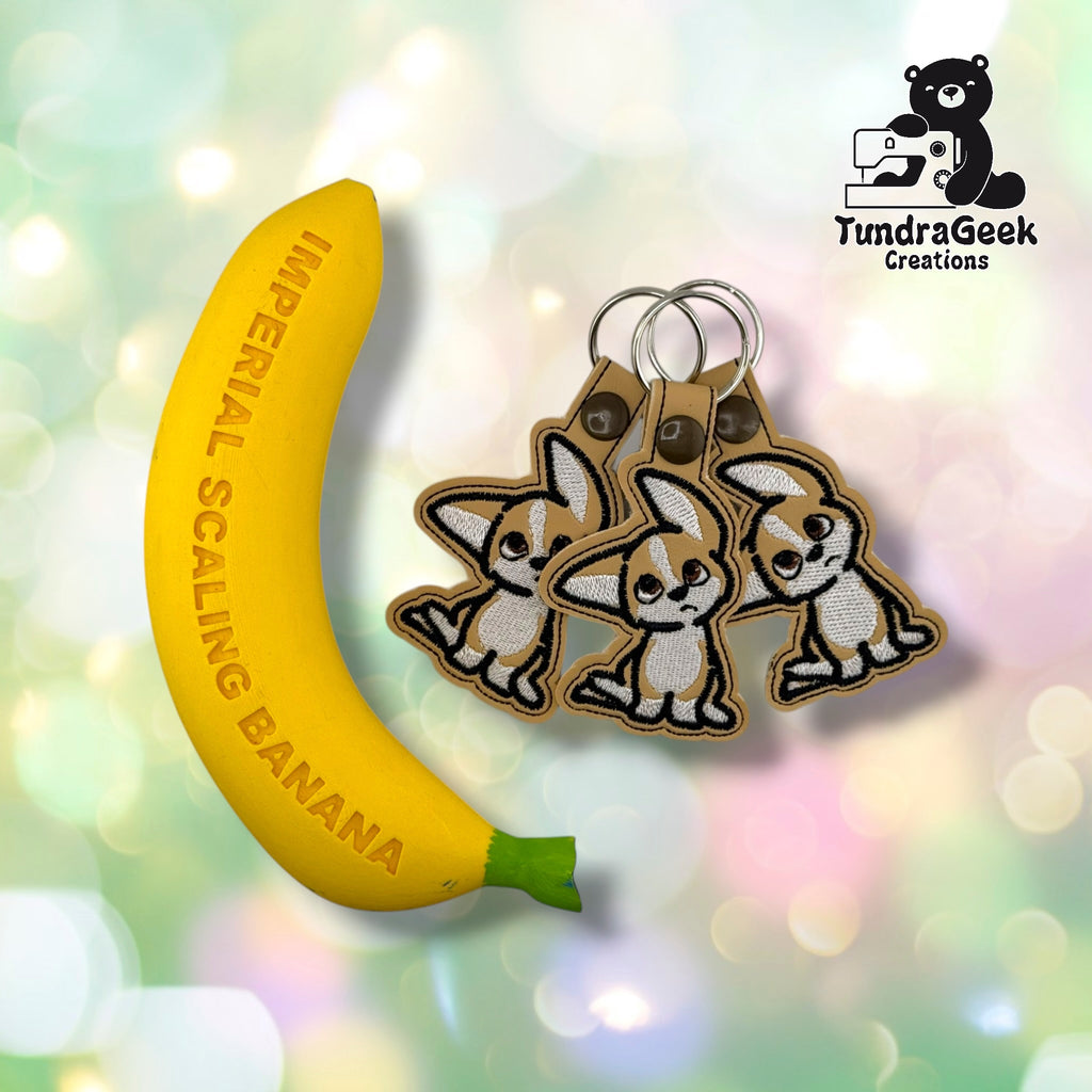 Three tan and white embroidered puppy keychains with metal rings attached to a yellow banana-shaped scaling tool. TundraGeek Creations logo visible in top right corner.