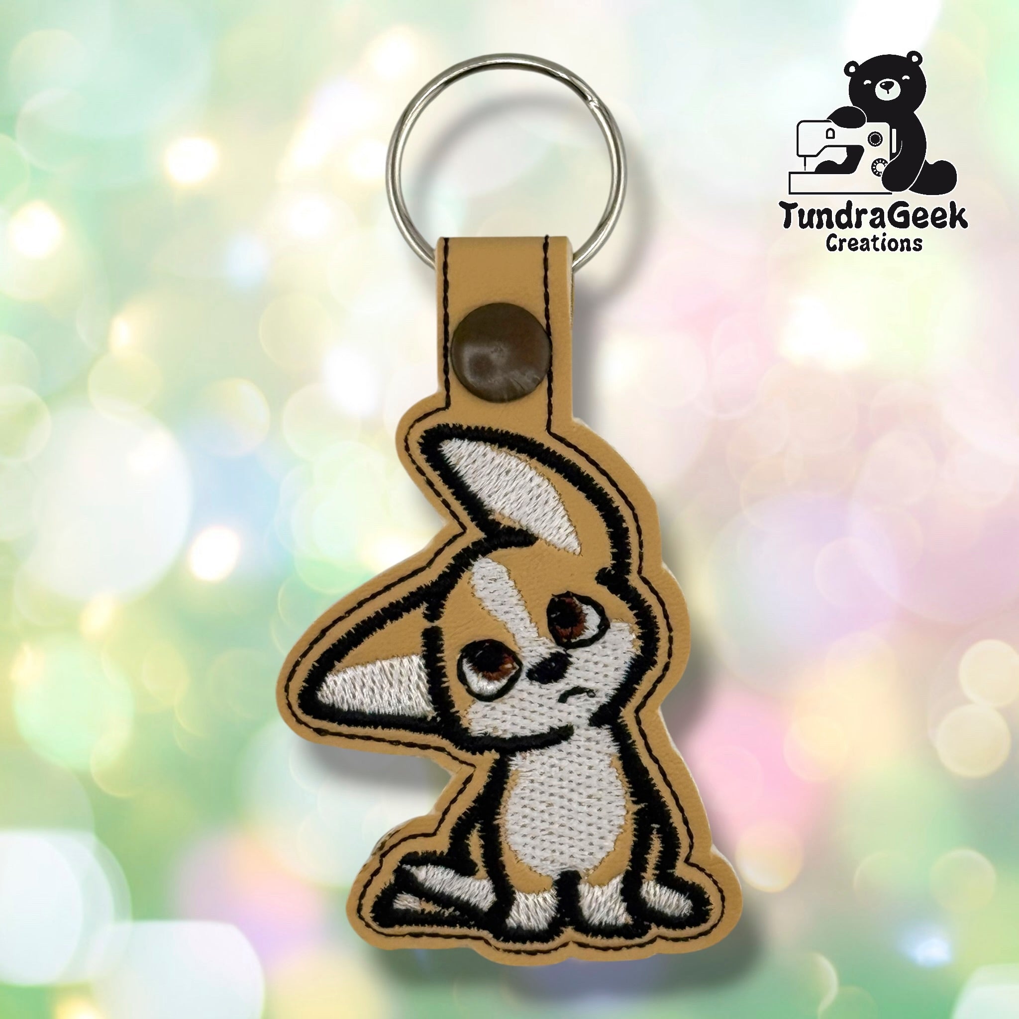 Tan embroidered keychain featuring a confused puppy with white and brown markings, attached to a metal keyring. TundraGeek Creations logo visible in top right corner.
