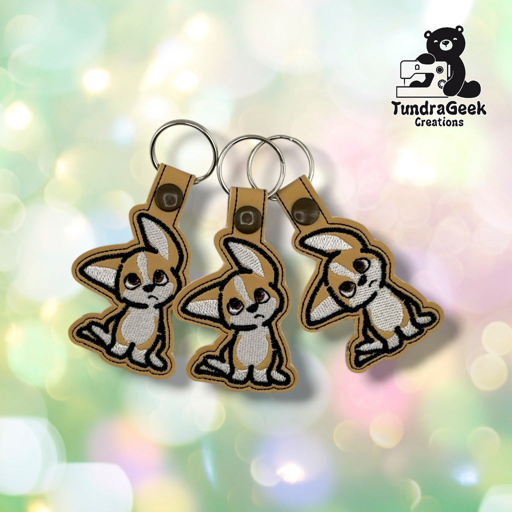 Three embroidered tan and white puppy keychains with confused expressions, attached to metal keyrings and clasps. TundraGeek Creations logo visible in top right corner.