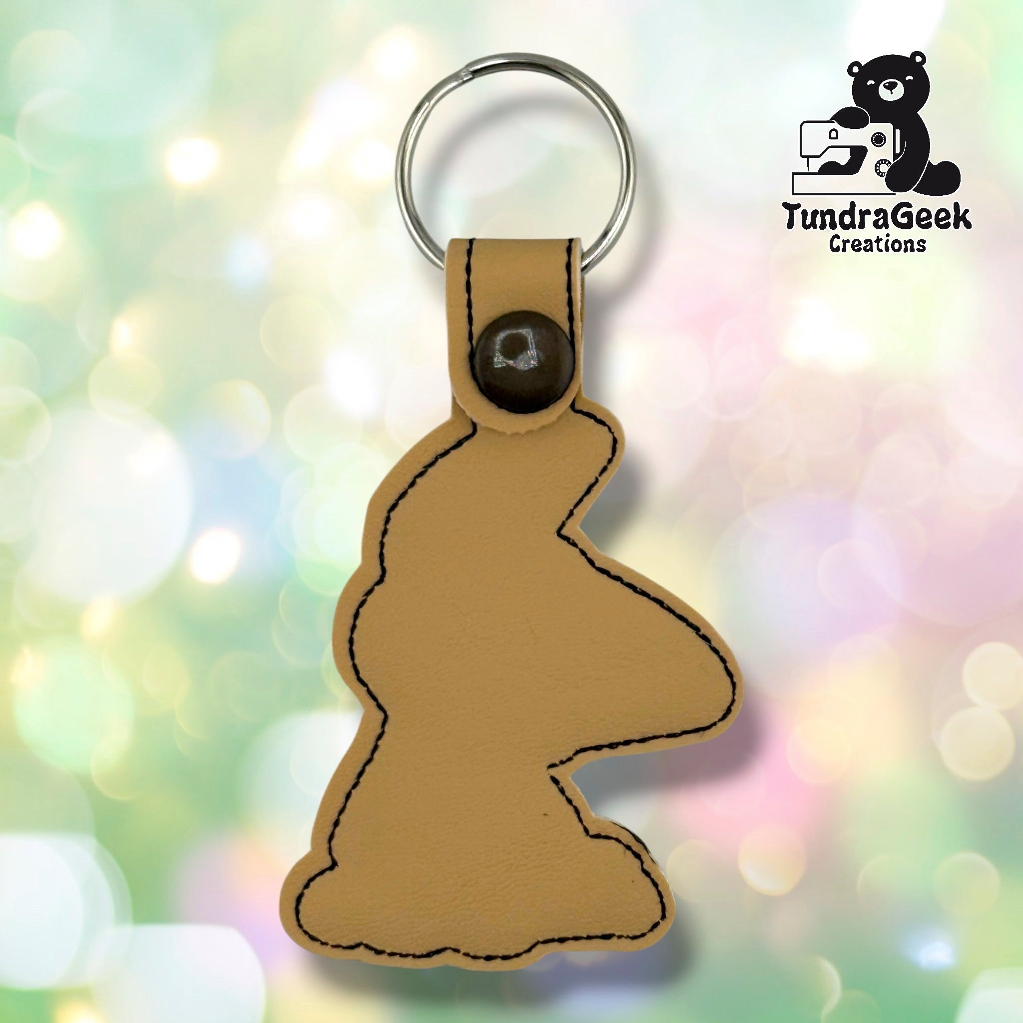 Tan vinyl embroidered keychain shaped like a confused puppy, attached to a metal keyring. TundraGeek Creations logo visible in the top right corner.