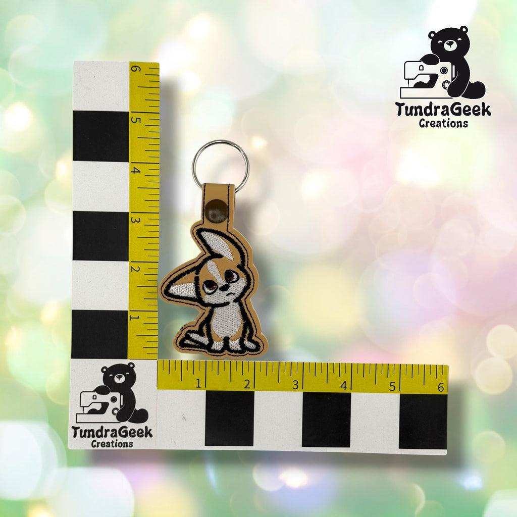 Tan embroidered keychain featuring a confused puppy with pointed ears, attached to a metal keyring. Measured against a ruler with TundraGeek Creations logo visible.