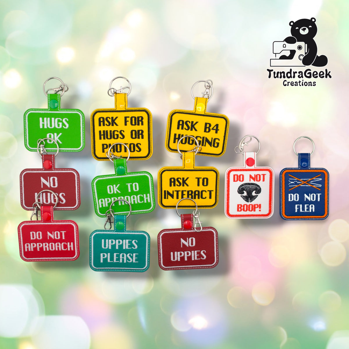 Eleven embroidered vinyl consent keychain badges on a soft bokeh background. Three overlapping columns of three: on the left, Hugs OK (green), No Hugs (dark red) and Do Not Approach (red); in the middle, Ask for Hugs or Photos (yellow), OK to Approach (green) and Uppies Please (teal); on the right, Ask B4 Hugging (yellow), Ask to Interact (yellow) and No Uppies (dark red). Two upright badges stand apart at the far right: Do Not Boop (white with red) and Do Not Flea (purple with orange).