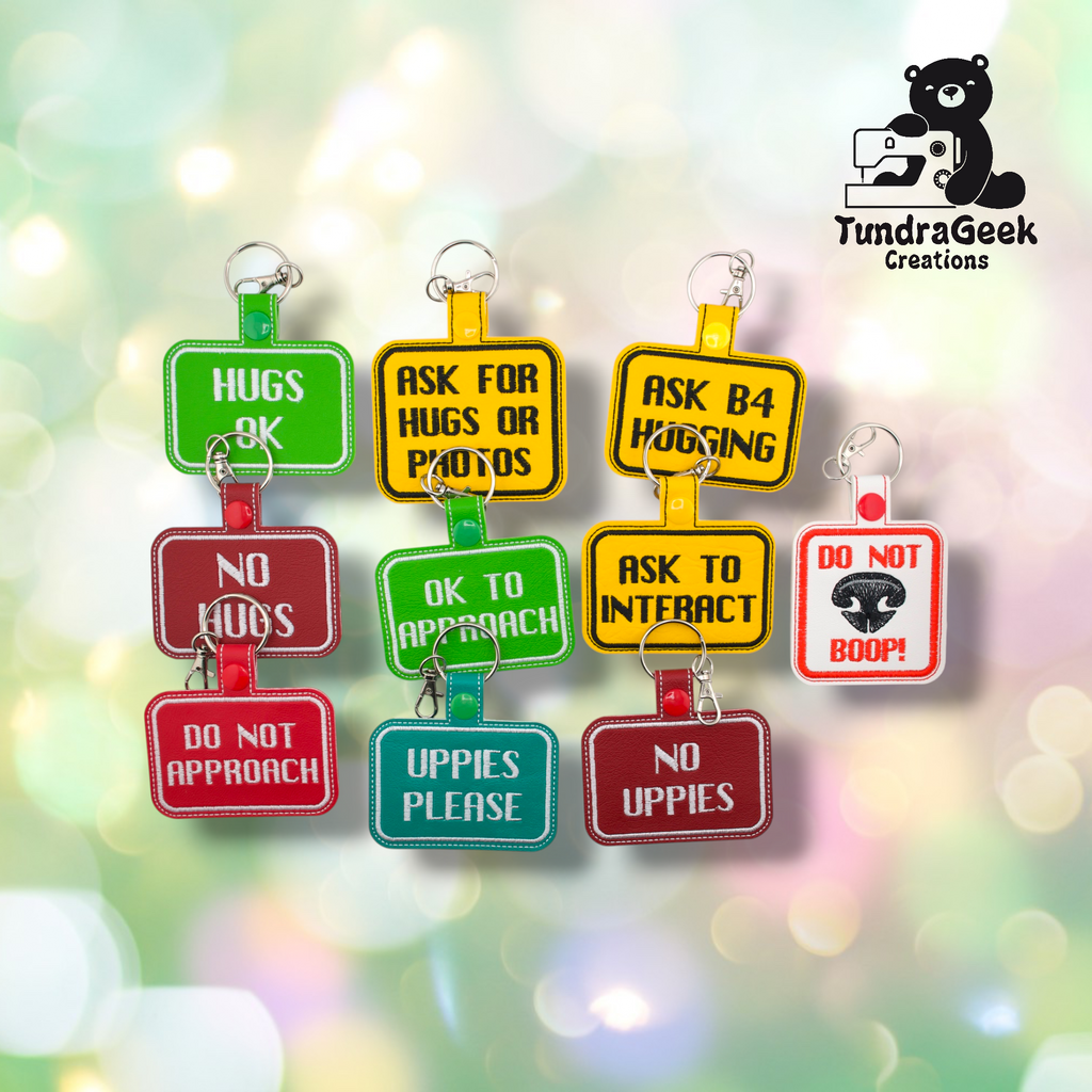 Nine embroidered vinyl consent keychain badges arranged in two rows on a soft bokeh background. Top row left to right: Hugs OK (green), Ask for Hugs or Photos (yellow), Ask B4 Hugging (yellow). Bottom row: No Hugs (red), OK to Approach (green), Ask to Interact (yellow), Do Not Boop (white/red). Front row: Do Not Approach (red), Uppies Please (teal), No Uppies (red).