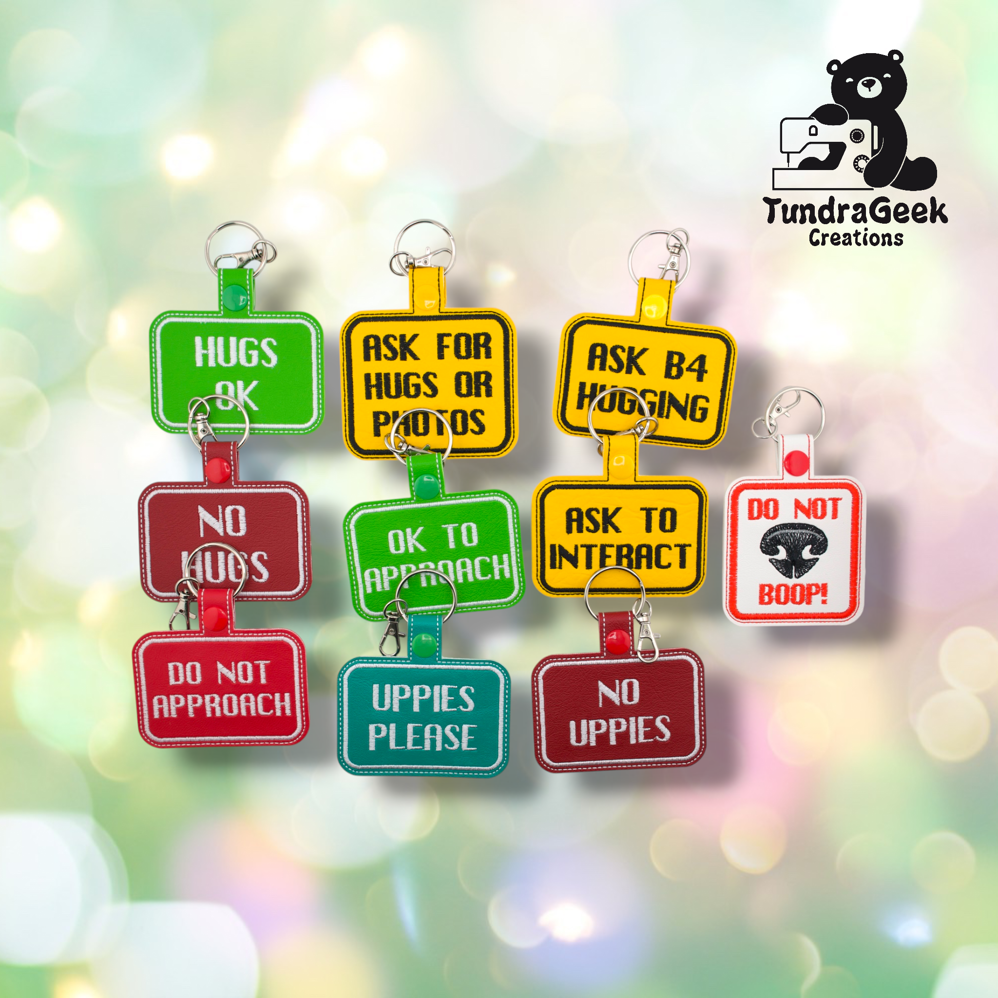 Nine embroidered vinyl consent keychain badges arranged in two rows on a soft bokeh background. Top row left to right: Hugs OK (green), Ask for Hugs or Photos (yellow), Ask B4 Hugging (yellow). Bottom row: No Hugs (red), OK to Approach (green), Ask to Interact (yellow), Do Not Boop (white/red). Front row: Do Not Approach (red), Uppies Please (teal), No Uppies (red).