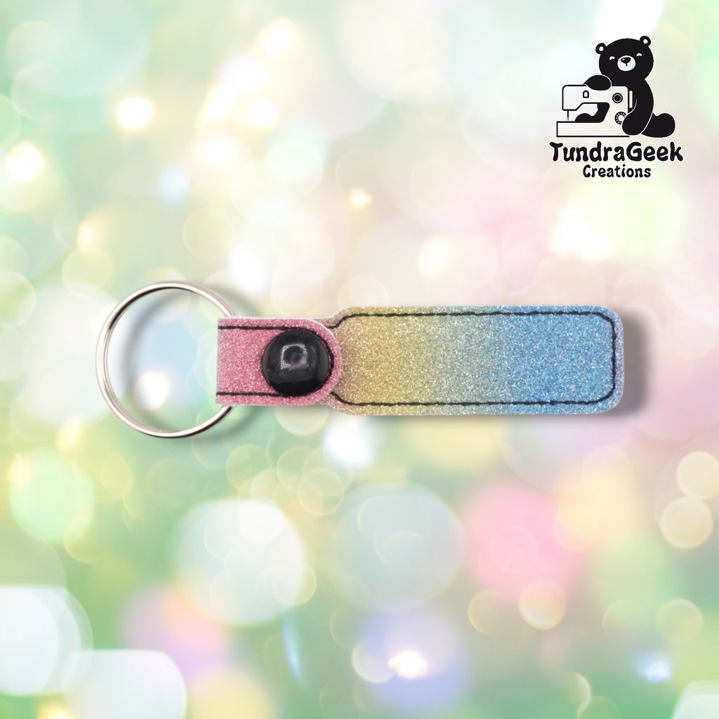 A glittery keychain with pink, gold, and blue gradient ombre finish, metal ring, and black button detail on soft pastel bokeh background.