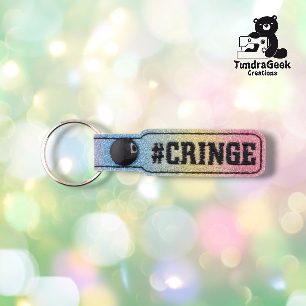 A glittery rainbow gradient keychain with "#CRINGE" embroidered text and a black snap button against a soft pastel bokeh background.