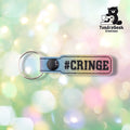 A glittery rainbow gradient keychain with "#CRINGE" embroidered text and a black snap button against a soft pastel bokeh background.