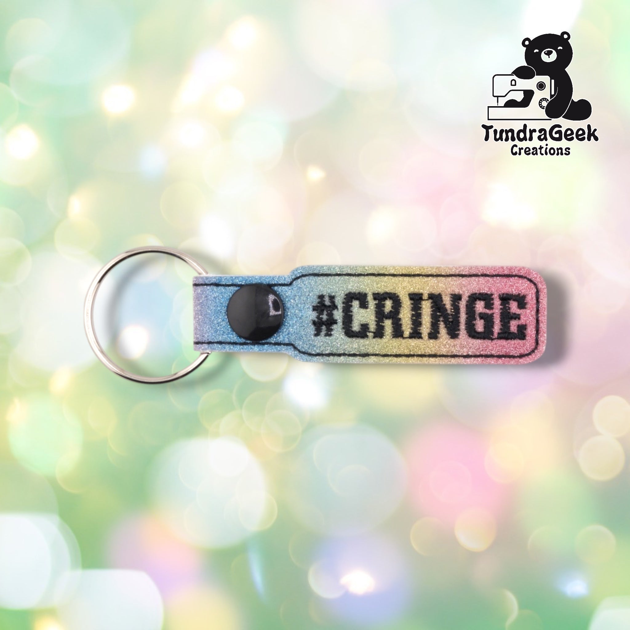 A glittery rainbow gradient keychain with "#CRINGE" embroidered text and a black snap button against a soft pastel bokeh background.