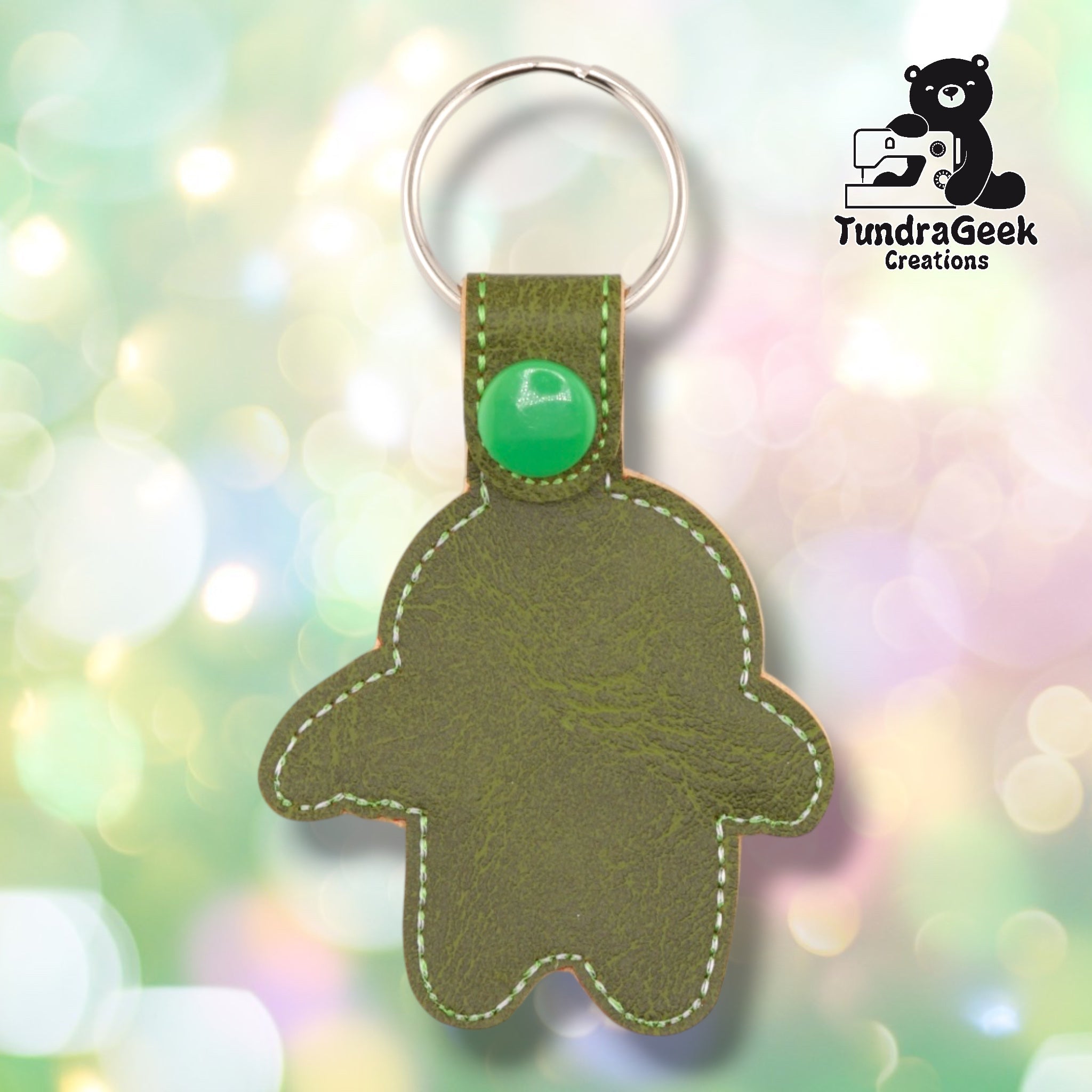 Olive green embroidered vinyl keychain shaped like a gingerbread person with white stitching and a green button detail, attached to a metal keyring. TundraGeek Creations logo visible in the top right corner.