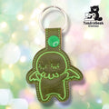 Front view of a single olive green vinyl keychain with a sleepy Cthulhu embroidered in lighter green outline detail, closed eyes, tentacles, and small wings, finished with a green plastic snap closure and split ring