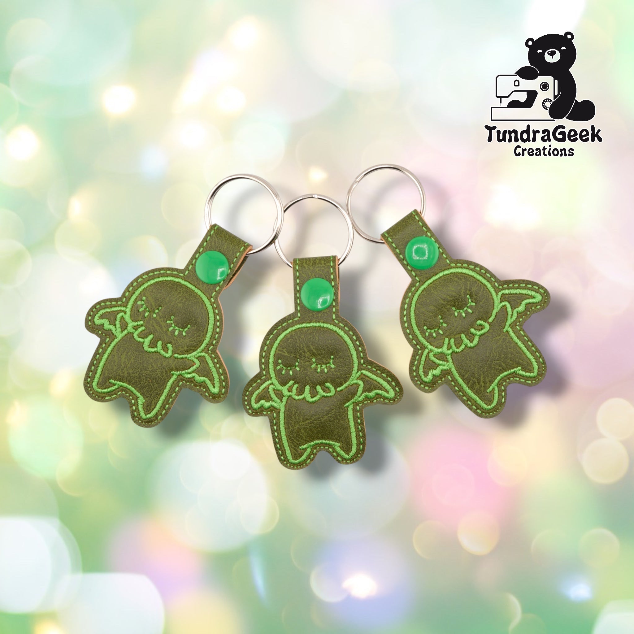 Three olive green embroidered Cthulhu keychains with closed eyes and tentacles, attached to metal rings. TundraGeek Creations logo visible in top right corner.