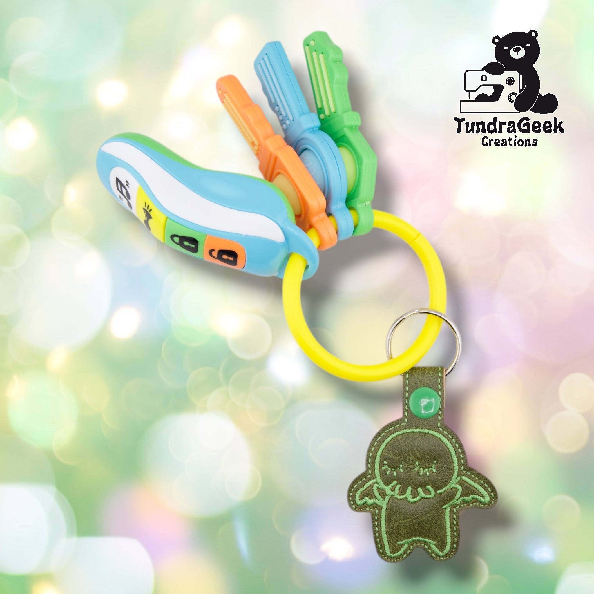 A colorful keychain with a yellow ring, blue and green toy keys and a green embroidered Cthulu charm against a soft bokeh background.