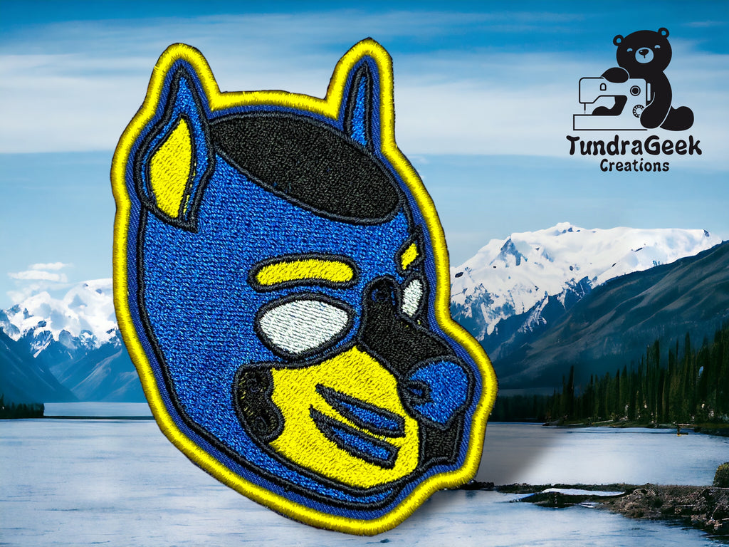 Close-up of a custom embroidered pup hood patch in blue and yellow with metallic thread accents, shown against a mountain and lake backdrop.